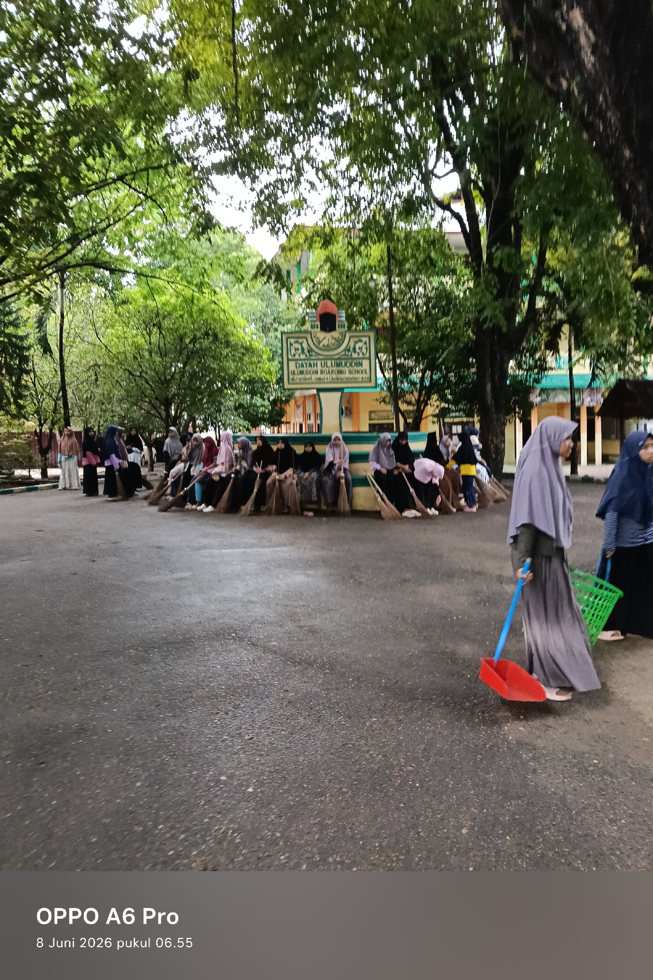 In Lhokseumawe, Indonesia, a group of students, likely female, are gathered outside a building identified by a sign as "DAYAH ULUMUDDIN ULUMUDDIN BOARDING SCHOOL". The students, predominantly wearing hijabs and modest clothing, are holding brooms. Some are sitting on a low wall or ledge, while others are standing. In the foreground, a woman wearing a grey hijab and skirt is walking with a red dustpan and a blue broom handle. To her right, a green plastic basket is visible. The scene takes place on an asphalt ground with scattered debris, suggesting a cleaning activity or a break. Lush green trees with dense foliage are visible in the background, creating a shaded environment. The building behind the students appears to be a school structure with yellow walls and green trim. The overall impression is one of an organized community activity, possibly a clean-up drive or a routine maintenance task within the school premises.