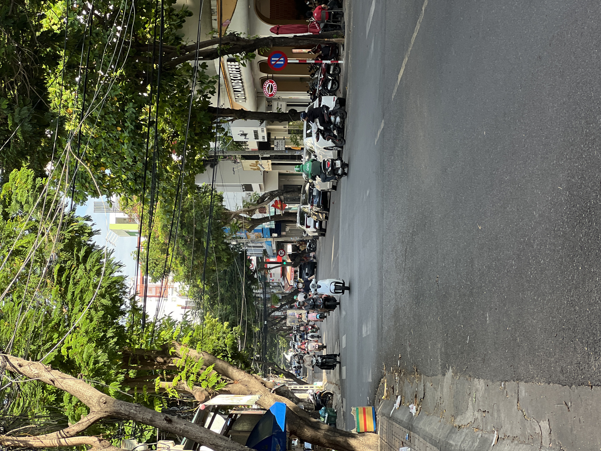This is a daytime, eye-level, outdoor, medium-shot photograph of a street scene in Nha Trang, Vietnam, likely in a commercial district. The street is paved and lined with trees whose branches, laden with green leaves, form a canopy overhead, with numerous electrical wires strung between them. The buildings along the street are a mix of commercial establishments, including one with a prominent sign for "HIGHLANDS COFFEE" featuring its logo. There are also other businesses with signage visible, such as "Ann."
The street is populated with numerous motorbikes, the primary mode of transportation. Several individuals are riding these motorbikes, some of whom are wearing helmets and face masks. One rider in a green uniform with "Grab" on his back is on a motorbike. There are also parked motorbikes along the side of the street. A few cars are visible, including a white car and a dark-colored sedan. People are seated at tables outside the "HIGHLANDS COFFEE" establishment.
The sidewalk on the left side of the street appears somewhat cluttered with debris and some trash cans. The lighting suggests it is either early morning or late afternoon, with sunlight casting shadows. The overall atmosphere depicts a busy urban street in Vietnam, with a mix of daily life and commercial activity.