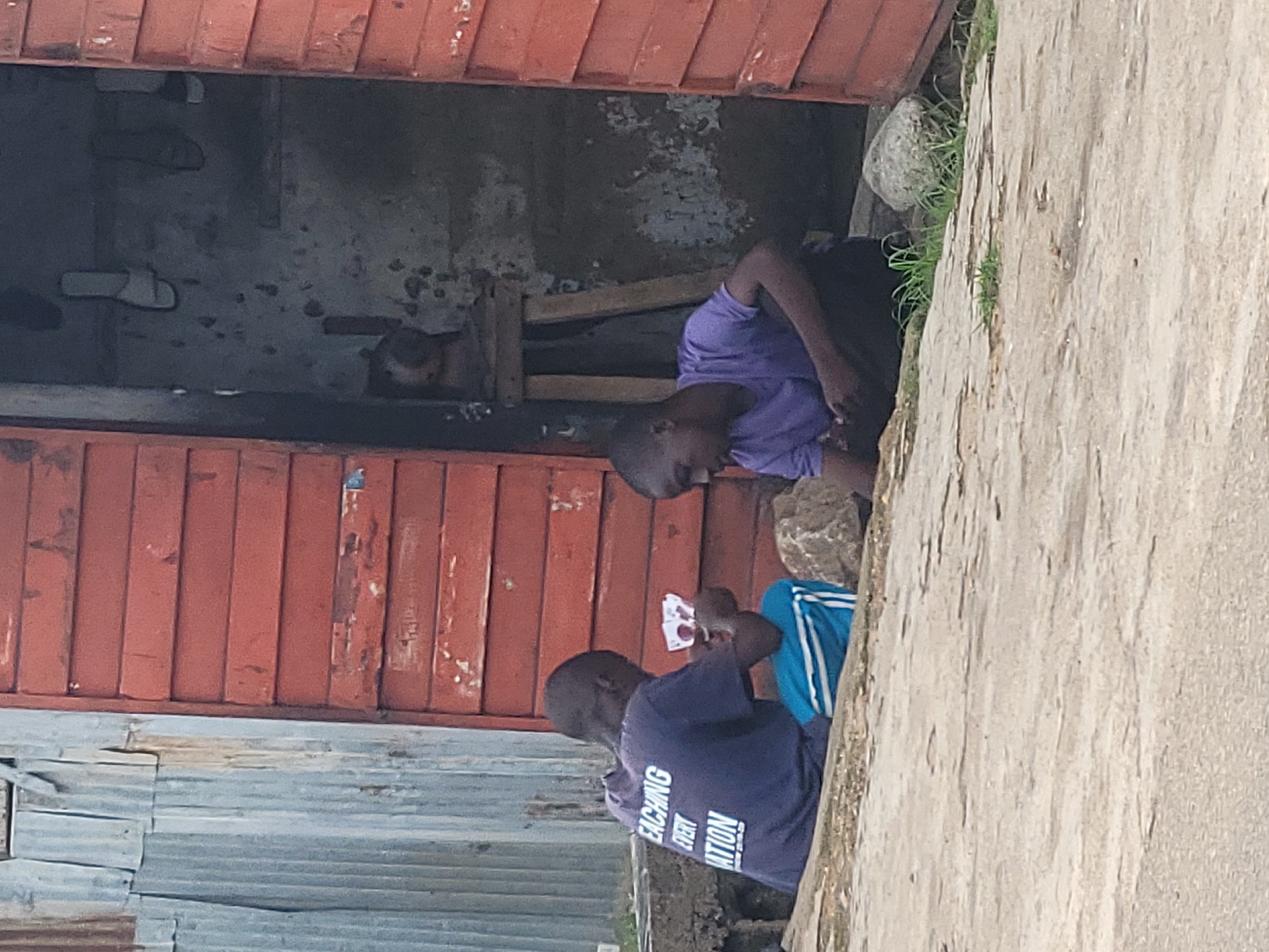 Two young males are seated on the ground in front of a rustic wooden structure. The structure has a red, weathered exterior and an open doorway revealing a dark interior. The male on the left wears a dark blue t-shirt with white lettering that reads "TEACHING EVERY NATION" and blue and white striped shorts. He is holding playing cards in his hands. The male on the right wears a purple t-shirt and dark trousers. He appears to be engaged with something on the ground in front of him. The ground is a rough, concrete surface with some patches of dry grass. To the left of the structure, there is corrugated metal sheeting. The scene suggests an outdoor setting in a somewhat impoverished urban environment. This scene is consistent with observations of daily life in informal settlements within the city of Jos, Nigeria, where such structures and outdoor activities are common.