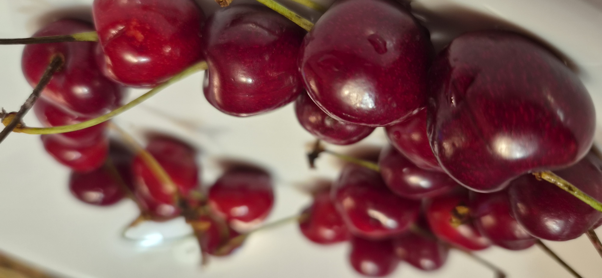This media file presents a close-up, still-life composition of several clusters of fresh cherries. The primary subjects are the deep red, almost burgundy-colored cherries, many of which are still attached to their green and brown stems.
The cherries appear ripe and plump, exhibiting a glossy, reflective skin that suggests freshness, possibly with a light sheen of moisture. They are arranged on a smooth, light-colored, reflective surface, which clearly mirrors their forms and vibrant color. The arrangement is somewhat scattered, with some cherries positioned closer to the viewer and others receding into the background.
A shallow depth of field is employed, bringing the cherries in the immediate foreground into sharp focus, while those further back in the composition are softly blurred, creating a pleasing bokeh effect. This focus draws attention to the rich texture and highlights on the individual fruits. There is no specific activity or event taking place; the image captures a moment of arranged produce.
Notable details include the contrasting vibrant red of the cherries against the crisp green of some stems, and the bright reflections on their surfaces. The lighting, possibly natural or controlled, enhances the cherries' natural sheen. There are no people, visible text, or explicit location cues within the image itself, though the context provided indicates the photo was taken in Varna, Bulgaria. The overall impression is one of natural beauty, freshness, and ripeness.