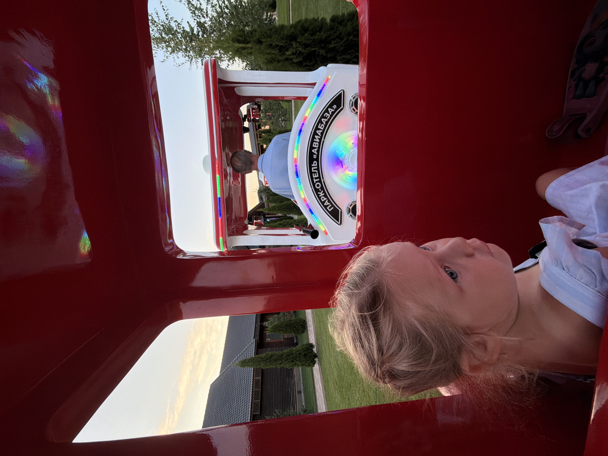 The image shows a young girl with blonde hair sitting in a red train-shaped ride. She is wearing a light blue garment. A man with gray hair, wearing a light blue collared shirt, is driving the train. The train has colorful lights and a sign that reads "ПАРК-ОТЕЛЬ «АВИАБАЗА»" (Park-Hotel AviaBaza). The background shows a lawn, trees, and a building with a dark roof. The scene appears to be at dusk, with a soft sky. This image was taken at the Park-Hotel AviaBaza in Kamyshin, Russia.
