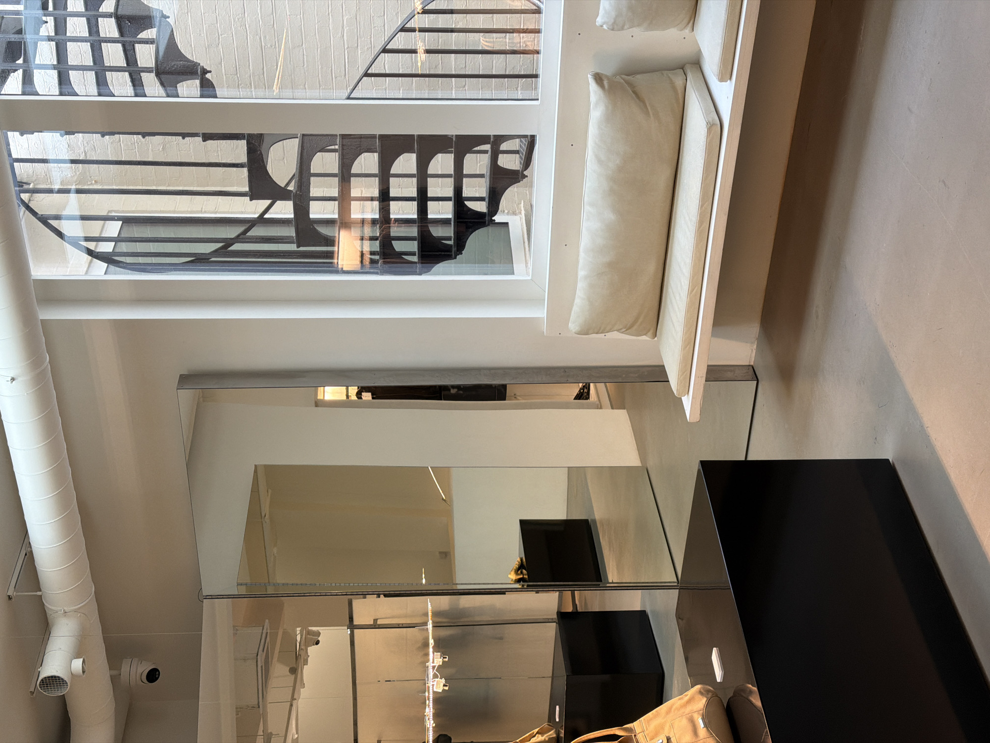 This media file captures an interior scene, likely a modern commercial or public space, shot from a slightly elevated perspective. The dominant feature is a large window on the left side, looking out onto a narrow exterior area. Through the window, a black metal spiral staircase with intricate detailing is visible against a light-colored brick wall, suggesting an urban setting like a courtyard or alleyway.
Inside, along the right side of the window, there is a minimalist white bench adorned with several off-white rectangular cushions, creating a clean and inviting seating arrangement. Below the window and stretching across the ceiling on the left, a large, white, ribbed air duct or ventilation system runs horizontally, with a white security camera mounted beneath it.
The foreground and bottom right of the image are dominated by highly reflective mirrored surfaces, which create complex reflections of the interior space. These mirrors reflect parts of the ceiling, the security camera, and another black rectangular object positioned further back in the room. A black, rectangular display or table stands prominently in the bottom right corner, and a light brown, possibly leather, bag or backpack is partially visible at its base.
The overall aesthetic is contemporary and clean, characterized by light colors, reflective surfaces, and functional elements. There are no people visible, and no specific activity is taking place. The lighting suggests it is daytime. No visible text is identifiable in the image.