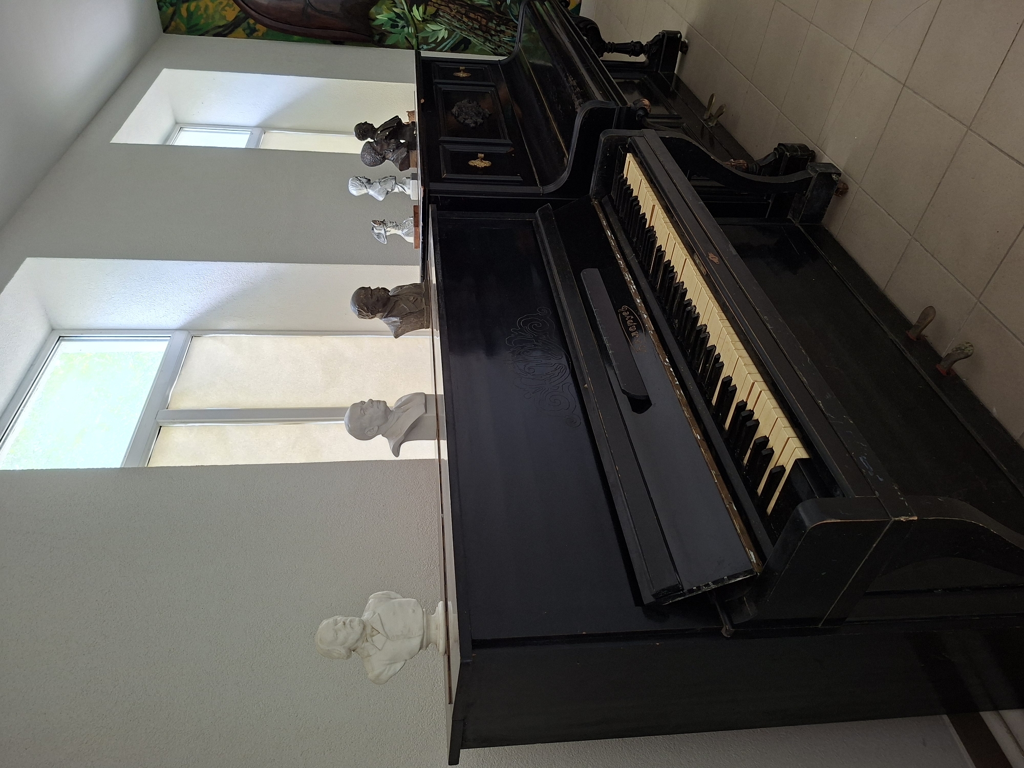 Two upright pianos, one on the left and one on the right, are positioned side-by-side in what appears to be a room with tiled flooring and tall windows. The piano on the left is black with visible ivory and black keys, and a decorative swirl pattern on its side. On top of this piano, there is a white bust of a man with a beard and a necktie. To the right of this bust, on the same piano, is a smaller white bust of a man. The piano on the right is also black and positioned slightly behind and to the right of the first piano. On top of the right piano, there are several small busts made of various materials, including bronze and white stone, depicting men in historical attire. Some of these busts are clustered together. The environment is brightly lit by natural light streaming in from the windows, which have blinds partially drawn. To the far right, a portion of a mural depicting trees and greenery is visible on the wall. The overall scene suggests a display of musical instruments and historical busts, possibly in a museum, cultural center, or private collection located in Ialoveni, Moldova.