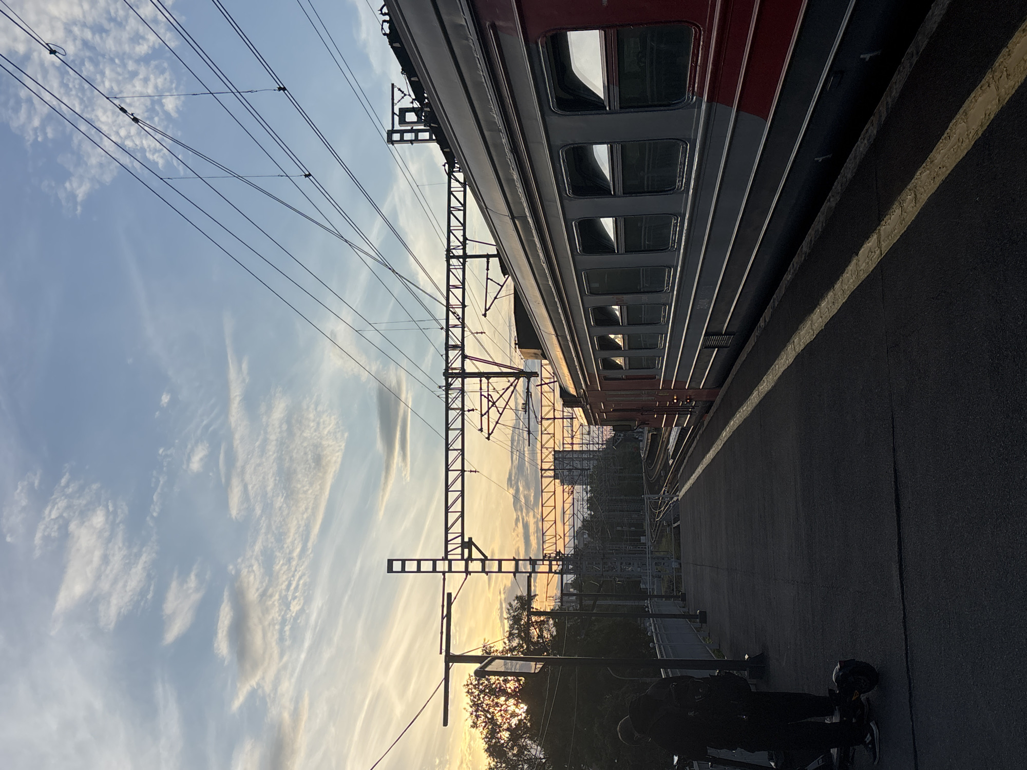 The image depicts a train station platform scene at either sunrise or sunset, viewed from a rotated perspective. When oriented correctly (landscape), a red and grey passenger train car is prominently positioned on the right, alongside a paved platform. The train appears stationary.
Above the platform and tracks, an intricate network of overhead electrical lines and robust metal support gantries stretches across the frame, silhouetted against the sky. The sky itself is a notable feature, displaying a blend of light blue and soft, wispy clouds, particularly illuminated with warm, golden hues near the horizon, indicative of the low sun. Trees and distant structures are visible further down the track, also silhouetted by the receding or rising light.
In the lower right corner of the platform, two individuals are partially visible in deep shadow, standing next to what appears to be a scooter or a similar personal transporter. They are largely obscured, making their exact actions unclear, but they seem to be waiting or passing through the station.
Notable details include the characteristic design of the train car, which is typical of an electric multiple unit or commuter train found in the region. The side of the train car has faded white text, possibly "C-80" or "C-B0". The warm, diffused light creates a serene and somewhat reflective atmosphere. The location is a railway station in Saint Petersburg, Russia.
