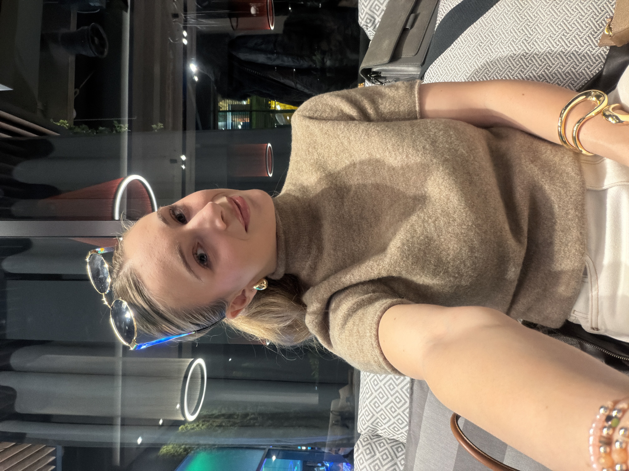 A young adult female is depicted in a close-up selfie, seated indoors at an establishment in Plovdiv, Bulgaria. She has fair skin, blonde hair pulled into a ponytail, and dark-lensed, gold-framed sunglasses positioned on her head. Her attire consists of a short-sleeved, light brown turtleneck sweater and white pants. Accessories include gold-toned hoop earrings and multiple bracelets on her right wrist, specifically a large, sculptural gold-toned cuff and several beaded strands. The subject maintains direct eye contact with the camera, exhibiting a slight head tilt and a subtle smile. She occupies a patterned upholstered bench, featuring a light gray fabric with a darker geometric design. The background, partially obscured by reflections in a large window, reveals interior architectural lighting, including circular ceiling fixtures and red pendant lamps. A dark jacket or coat is visible hanging on a fixture. Blurred exterior lights are also visible through the window, indicating evening or night conditions.
