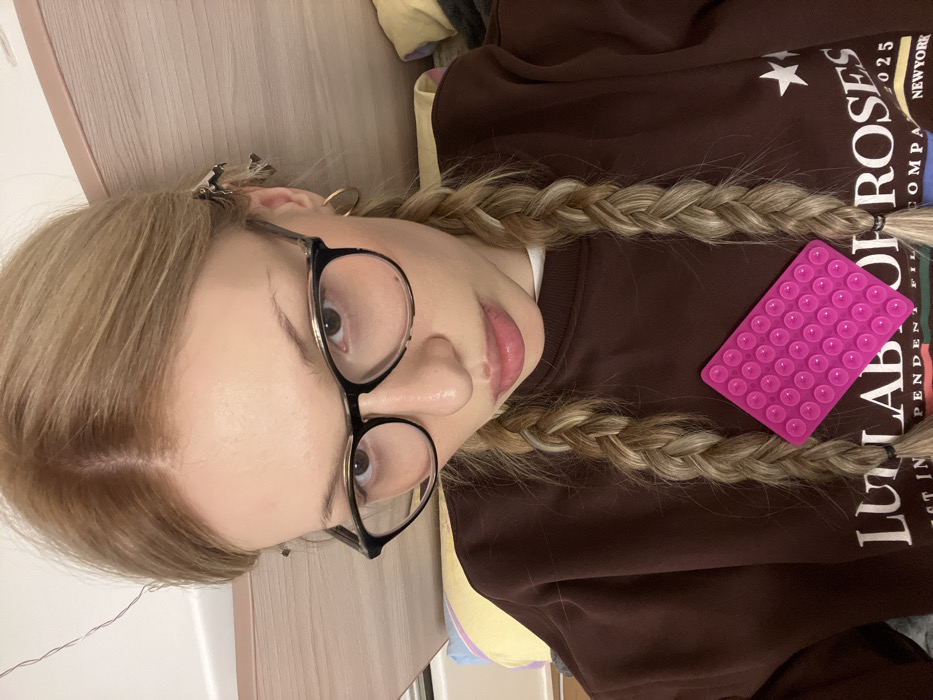 The image displays a close-up, rotated portrait of a person, likely a young woman, taken from an overhead perspective. The image is oriented such that the person's head is on the left side of the frame and their body extends to the right.
The person has long, blonde hair styled in two thick braids that fall over their shoulders and onto their chest. The hair roots show a slightly darker, natural blonde or light brown color. They are wearing a pair of eyeglasses with dark, possibly black or tortoiseshell, frames that have a slight upward flick at the outer corners. Their eyes, visible through the glasses, are light brown or hazel. Their lips are slightly parted and appear to be adorned with a glossy finish. On the left side of their head (top-right in the image), a few silver hair clips are visible, holding back some strands. A simple gold hoop earring can be seen in their left ear.
The person is wearing a dark brown long-sleeved sweatshirt or sweater. A white collar or neckline of an undershirt is visible beneath the brown garment. On the front of the brown sweatshirt, there is white text and a star graphic. Partially visible words include "NEW YORK 2023", "LAB OF ROSES", "COMPANY", "PENDENT", and "LULU".
Prominently placed on the chest of the sweatshirt, between the two braids, is a bright pink rectangular silicone mat. This mat is covered uniformly with numerous small, circular suction cups.
The background appears to be a light-colored, possibly wooden or painted wall surface, visible behind the person's head and to the left. Some soft, light-colored fabric or clothing is visible in the top right corner of the frame. The lighting is soft and even, suggesting an indoor setting. The overall impression is a casual, self-portrait style photograph.