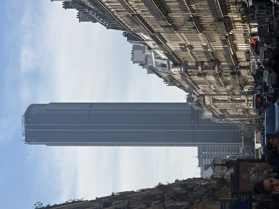 The image displays an urban street scene in Paris, France, looking south along Rue de Rennes towards the Tour Montparnasse. The Tour Montparnasse, a tall, dark, rectangular skyscraper with a slightly rounded top, stands prominently in the background against a light blue sky with scattered white clouds. On either side of the street, multi-story Haussmannian buildings line the thoroughfare, characterized by light-colored stone facades, numerous windows, and ornate wrought-iron balconies. Commercial establishments occupy the ground floors of these buildings. The foreground shows a busy street with vehicular traffic, including multiple cars and a city bus. Pedestrians are visible on the sidewalks. A street sign and an information panel are present on the left side of the street. One individual with dark hair is visible in the lower left corner, holding a mobile device near her ear. The scene is illuminated by bright daylight.