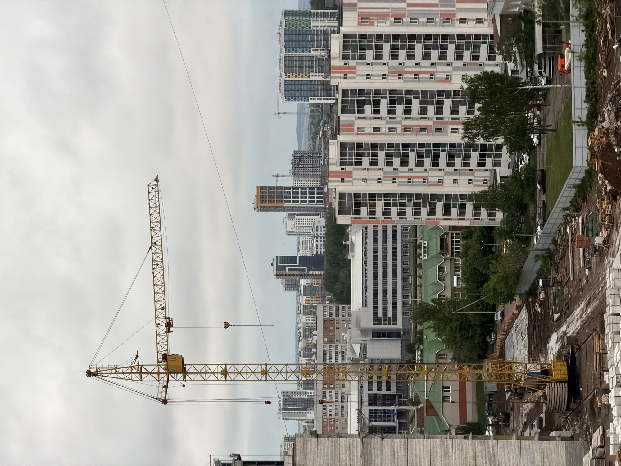 The image shows a construction site in Naberezhnye Chelny, Russia. A large yellow crane dominates the foreground. Beyond the construction site are multiple high-rise buildings, forming a dense urban landscape. The sky is overcast, suggesting a cloudy day. The construction site appears to be active, with materials and equipment visible.