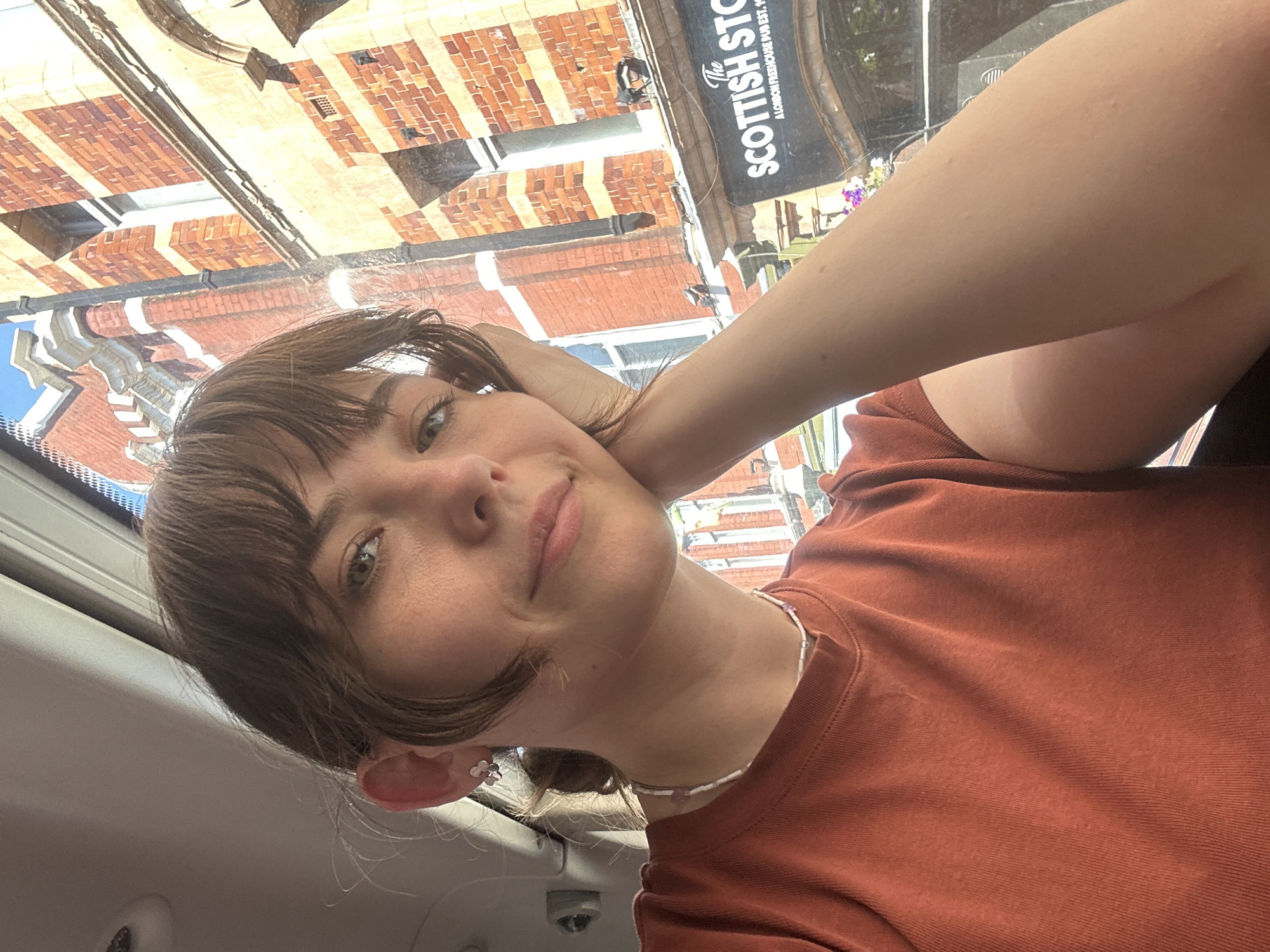 A young woman with brown hair and bangs is captured in a selfie. She is wearing an orange t-shirt and a beaded necklace. Her left hand rests on her cheek, and her gaze is directed towards the camera. Behind her, a brick building with white trim is visible through a window. A sign on the building reads "The Scottish Ston" with the text "A LONDON FREEHOUSE PUB EST. 19" visible below it, suggesting the location is The Scottish Ston, a pub in London. The sky appears blue and the scene is illuminated by sunlight, indicating it is daytime.