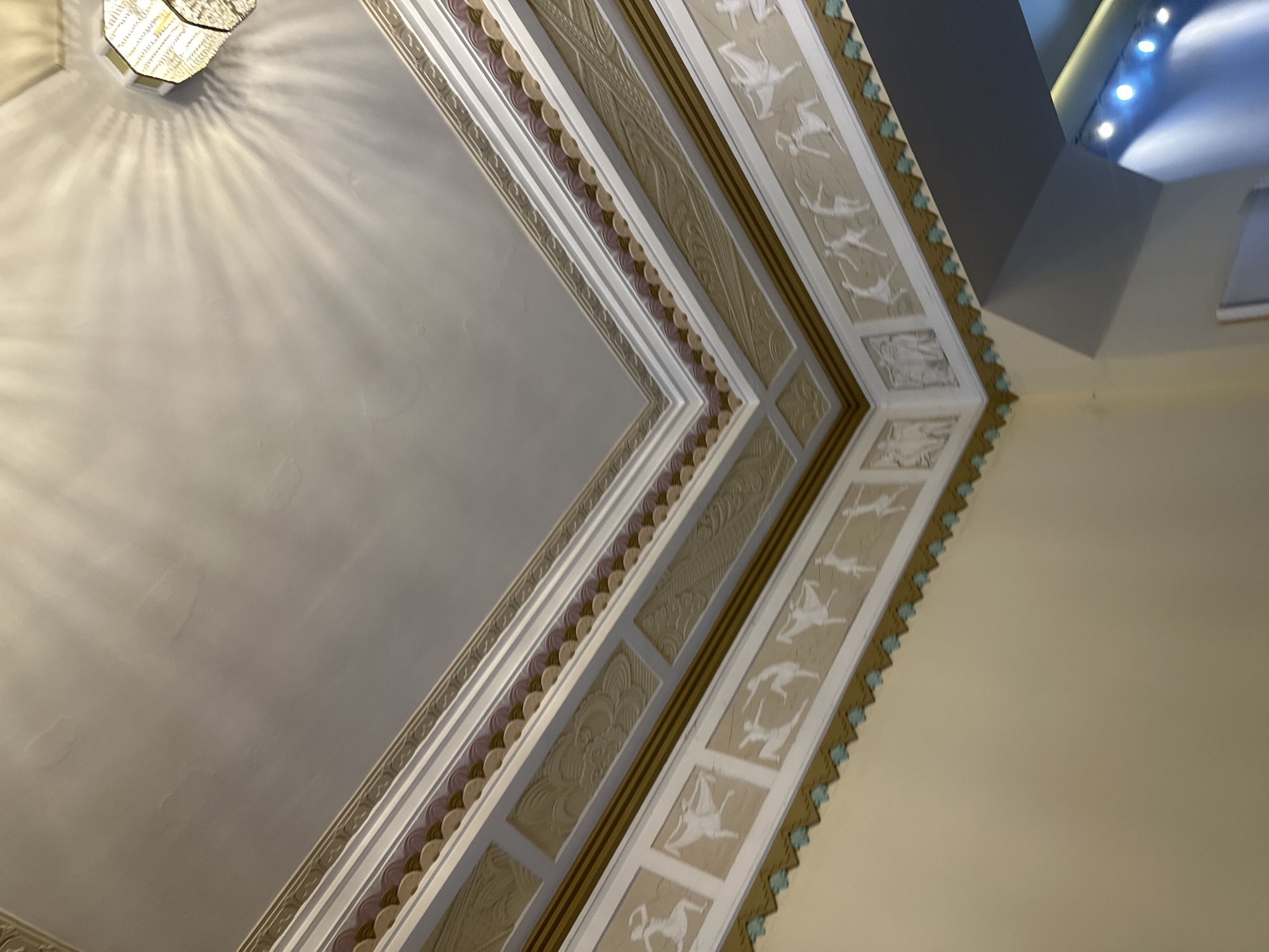 The image depicts a portion of a ceiling with ornate decorative moldings. The moldings feature various patterns, including geometric shapes, stylized floral designs, and a row of figures (possibly depictions of humans or animals). The ceiling is illuminated by a light fixture that casts radial shadows across the surface. The colors are predominantly neutral, with shades of white, beige, and gold. The location is Unknown, Unknown.