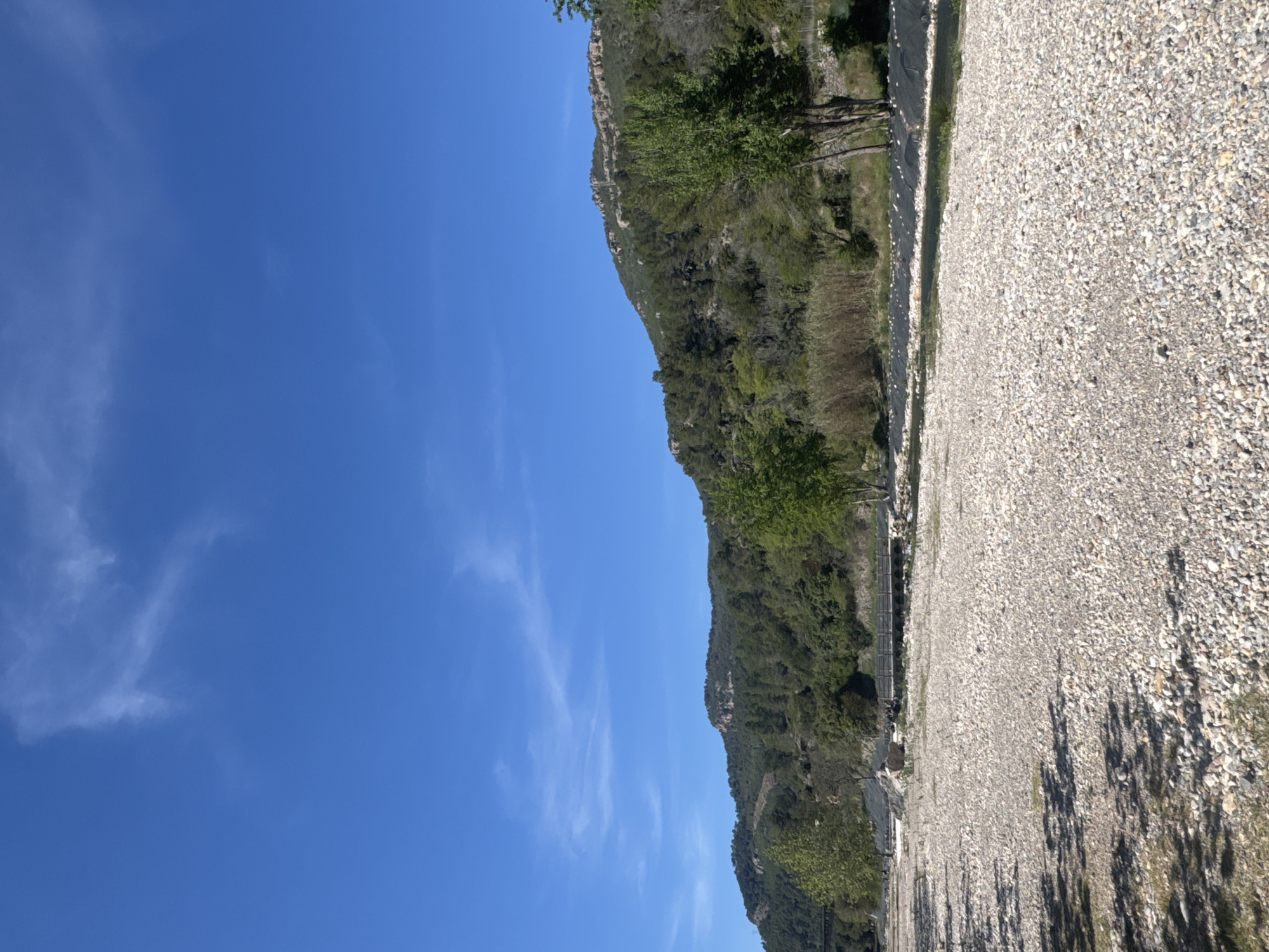 The image depicts a landscape in Montanejos, Spain. A rugged, green-forested hillside slopes downwards from the upper-middle section of the frame. To the right of the hillside is an extensive gravel plain composed of small, light-colored rocks. Tall shadows are cast across the gravel. To the left of the hill is a clear blue sky with a few scattered wisps of white clouds. A metal fence runs along the transition from the gravel plain to the hillside.