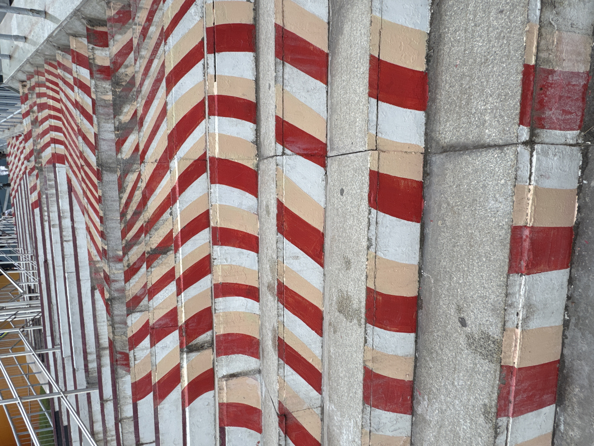 A set of concrete stairs, likely located in Ufa, Russia, are painted with alternating red and beige stripes on their risers. The stairs are angled, with a metal railing visible on the left side, suggesting they are part of a public walkway or entrance. The concrete is weathered, with some scuff marks and stains. No people are visible in the immediate foreground, though figures can be seen ascending the stairs in the background. The lighting suggests it is daytime.