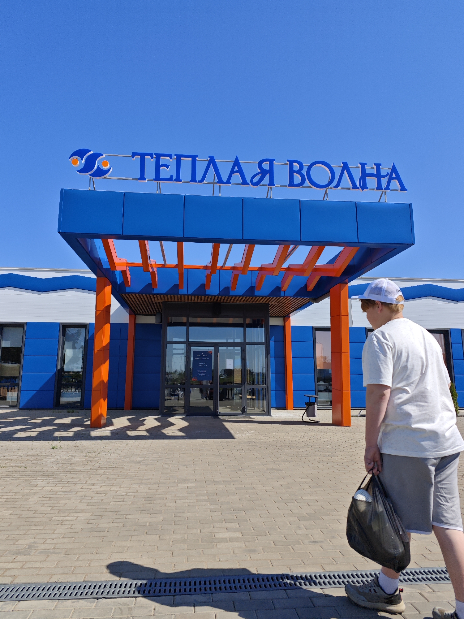 A full shot captures a male figure walking right in the foreground, near the "Teplyy Volna" building located in Votkinsk, Russia. The man wears a white t-shirt, grey shorts, sneakers, and a white cap. He carries a black bag.
The building's entrance features a blue and orange canopy. The sign above the entrance reads "TEПЛАЯ ВОЛНА" in Cyrillic. The building's facade alternates between blue panels and white wavy lines. A glass entrance door is visible with signage posted. A drainage system runs across the paved area in the foreground. The sky is clear and blue.