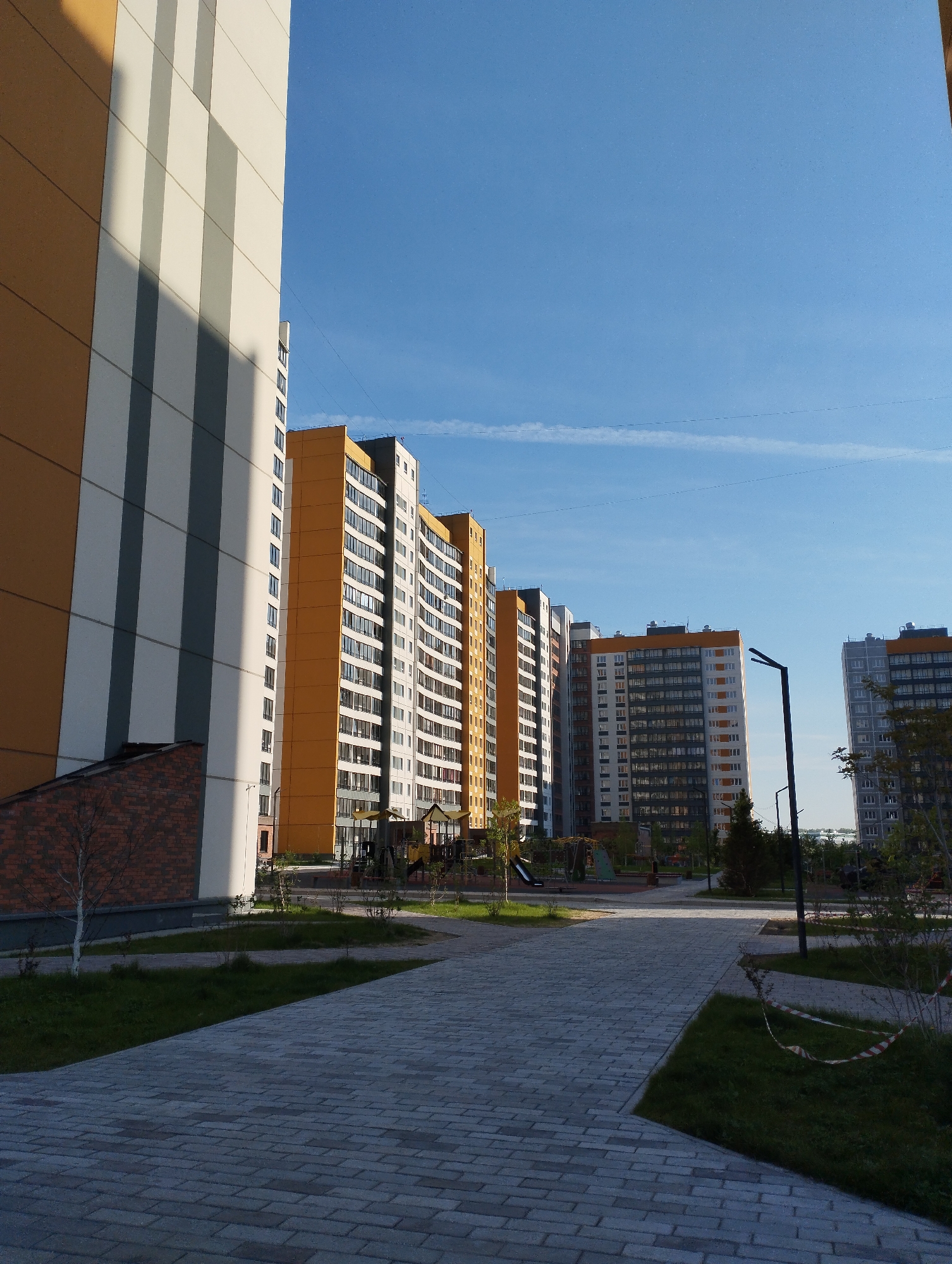 The image depicts a courtyard within the residential complex "Vesna" in Sadovyy, Russia. The foreground shows a paved walkway constructed of gray brick pavers. Adjacent to the walkway are patches of green grass and young trees. Buildings of varying heights are visible, featuring facades in shades of white, gray, and orange. The buildings are of modern design with multiple windows. In the center of the courtyard, there is a playground with slides and climbing structures. The sky is blue and mostly clear. A street light is partially visible in the right of the image. Red and white striped tape is around the grass, likely marking off an area.