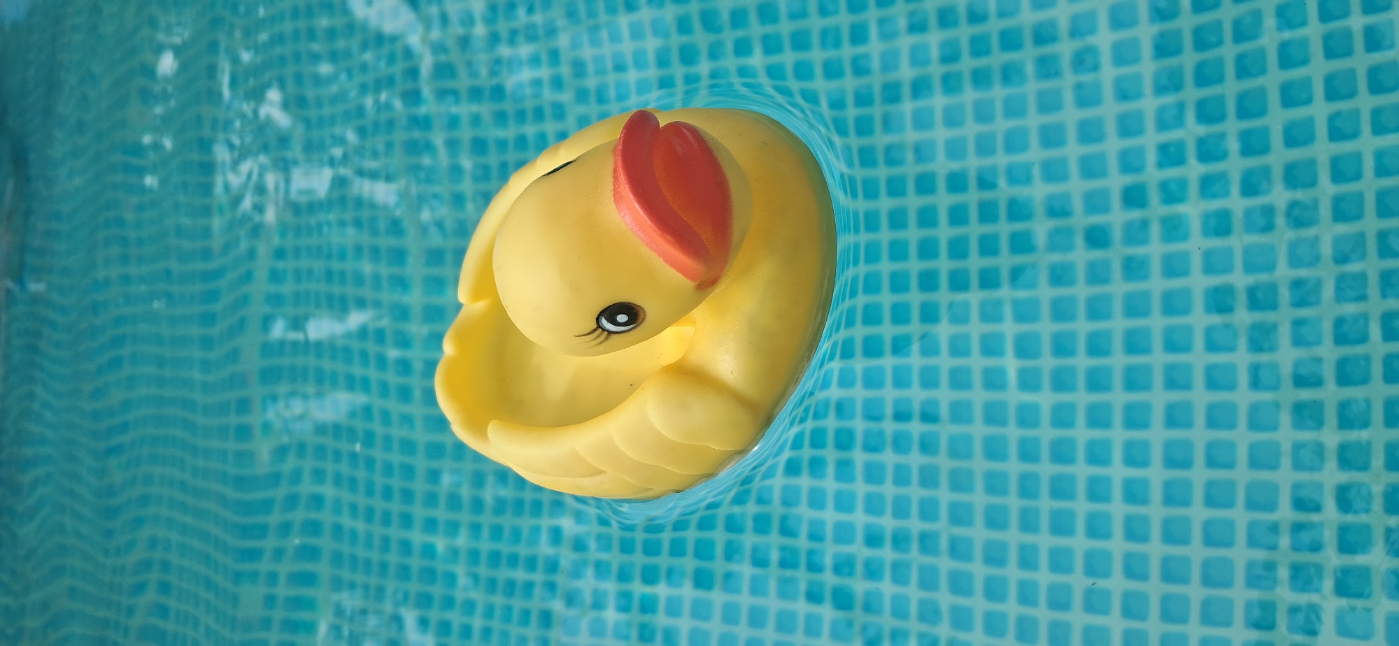 A yellow rubber duck floats on the surface of a swimming pool. The pool water is a vibrant turquoise and the bottom is tiled with a grid of light blue squares. The duck is positioned slightly off-center in the frame, with its body curved and its head turned to the right. Its beak is orange, and it has a black eye with a white highlight and a short, dark eyelash on its left side. The duck appears to be casting a subtle shadow on the pool floor. The image captures the playful, summery atmosphere of a day spent by the pool. The location is not discernible from the image.