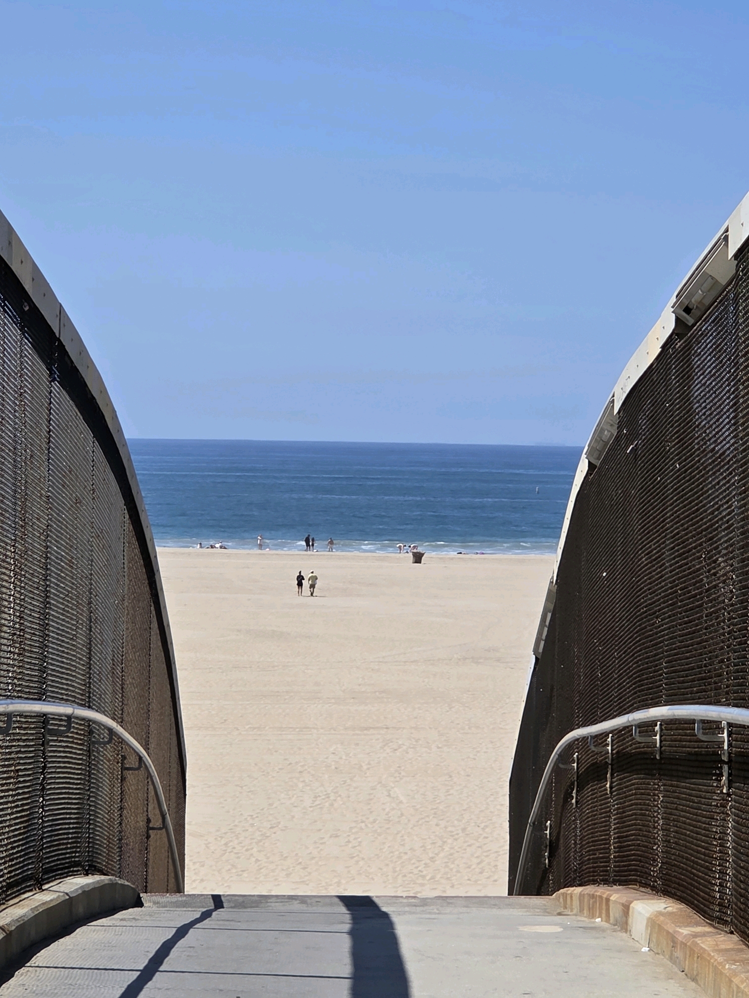 The image shows a view of the Santa Monica State Beach in Santa Monica, California, United States of America. The perspective is from the top of a pedestrian overpass that leads down to the beach. The overpass has metal mesh walls and a metal handrail. The beach is wide and sandy, with the Pacific Ocean in the background. The water is a deep blue, with gentle waves breaking on the shore. Several people are visible on the beach and in the water, engaged in various activities such as sunbathing, walking, and playing. A trash receptacle is also visible on the sand. The sky is clear and blue. The lighting suggests it is daytime, likely mid-morning or afternoon, with bright sunlight casting shadows.