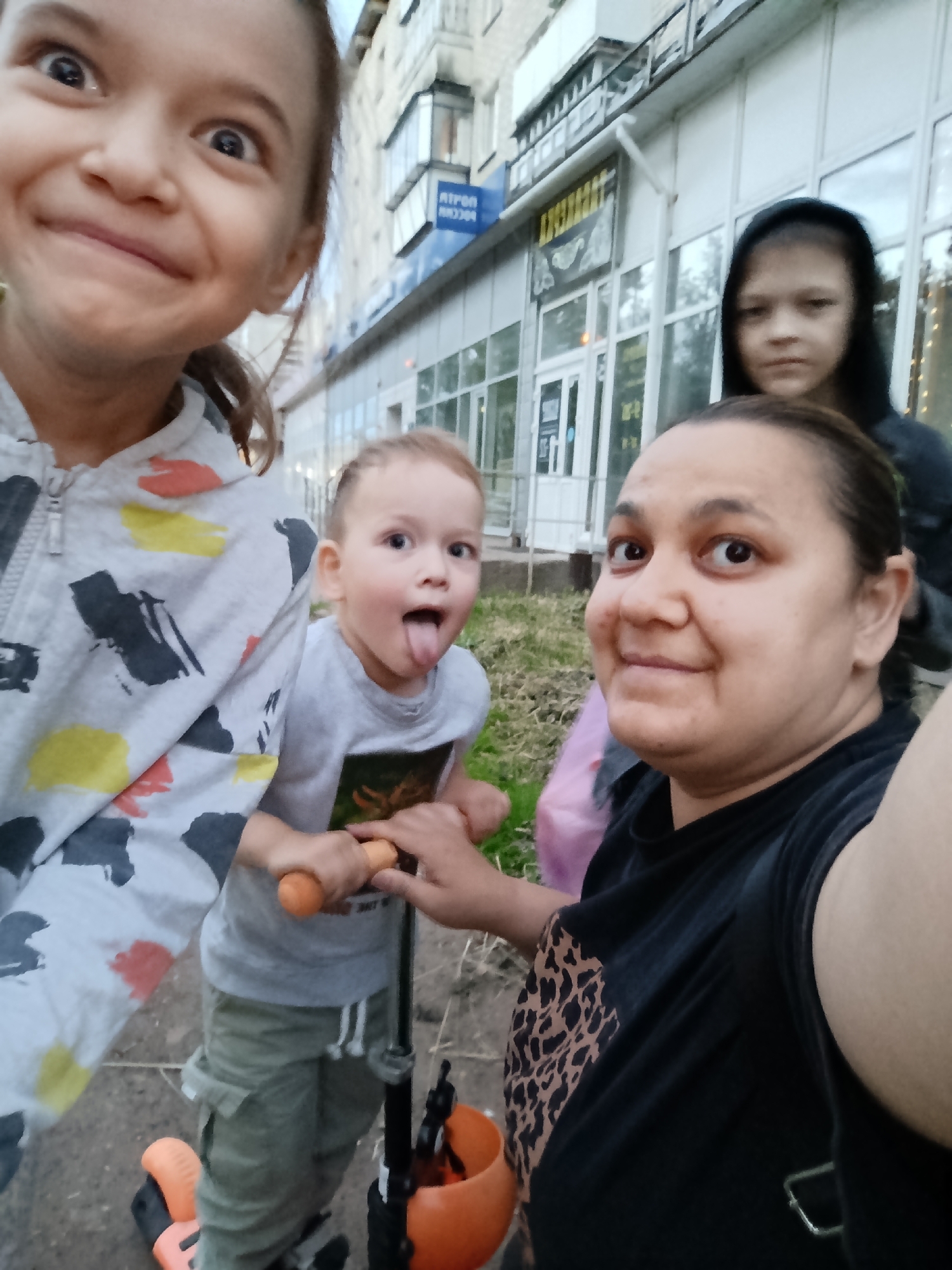 This is a medium shot, taken from a low angle, of a group of four people on a sidewalk in front of a building. The primary subject is a woman in the foreground, looking at the camera and smiling. She is wearing a black t-shirt with a leopard print design on the left side. To her left and slightly behind her are two children. The child on the immediate left, in the foreground, is a young girl making a funny face at the camera with her eyes wide and tongue sticking out. She is wearing a gray hooded jacket with abstract colorful shapes. To her right and further back, a younger child, a boy, is also sticking his tongue out playfully. He is wearing a gray t-shirt and khaki pants, and is holding the handlebars of an orange and black scooter. In the background, behind the woman and slightly to the right, is another child, a girl, wearing a black hooded sweatshirt, looking off to the side with a neutral expression. The setting appears to be a street in Chaikovsky, Russia, with a building in the background that has storefronts and windows. One storefront has a sign with Cyrillic text, which appears to read "РОСТ". Another sign above that has "РИПОЛ" visible. The lighting suggests it is daytime, possibly late afternoon. The overall mood of the image is playful and familial, with the children displaying energetic and humorous expressions.
