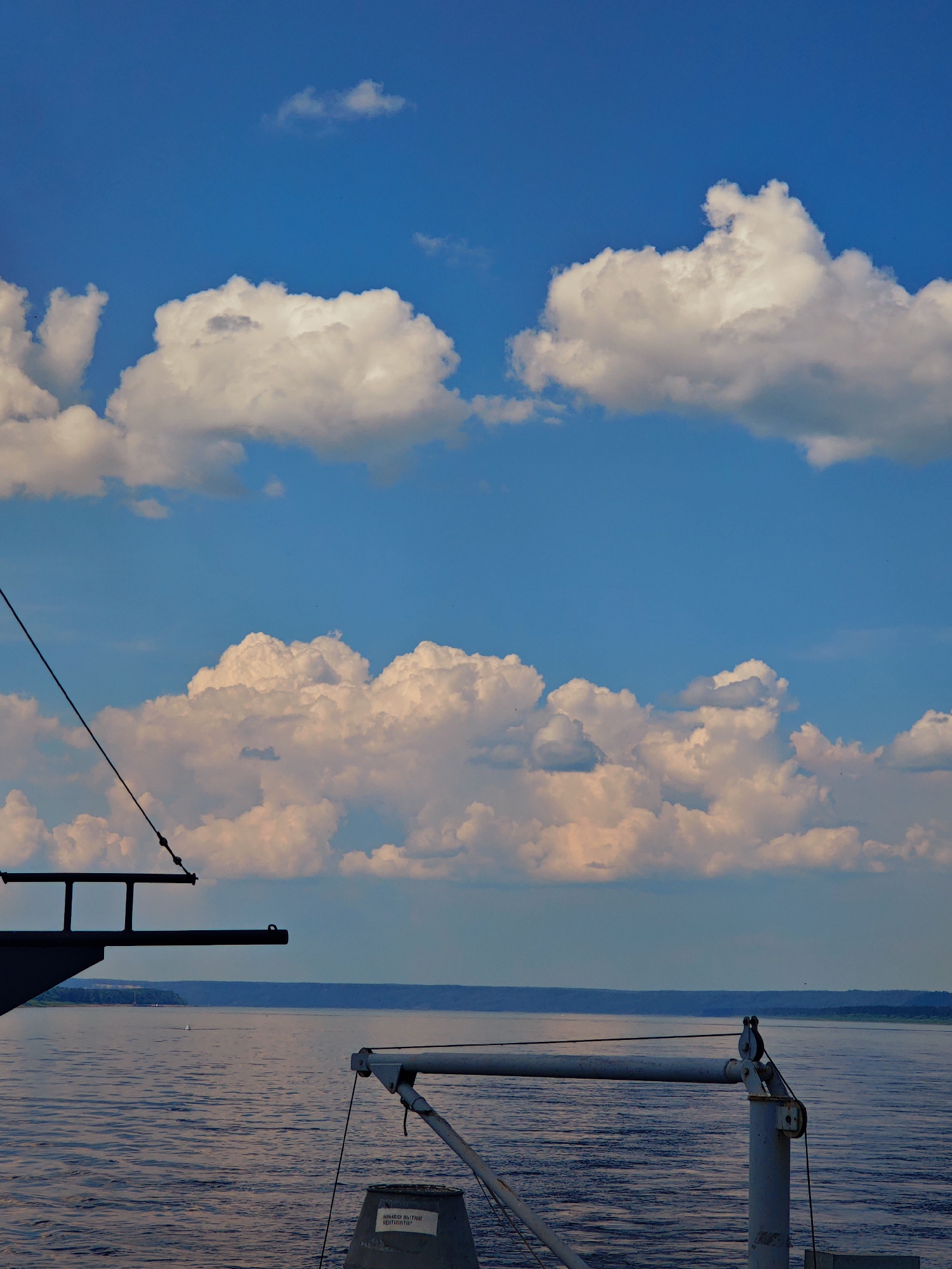 The image depicts a body of water, likely a river, under a partly cloudy sky. The water surface exhibits gentle ripples, reflecting the light of the sky. In the foreground, a portion of a vessel is visible, including metallic structures and rigging. A darker, angled structure on the left appears to be part of the ship's deck or superstructure. To the right, a lighter colored, angled arm with pulleys and cables extends into the frame. A cylindrical object, possibly a storage bin or equipment housing, is situated at the bottom right, bearing some white text. In the mid-ground, the opposite shore is visible as a dark, tree-lined expanse under a hazy sky. The overall scene suggests a calm day on the water, possibly during daylight hours given the brightness of the sky and clouds.
There is no information in the image to suggest the location is the city of Podkamennaya Tunguska, Russia.