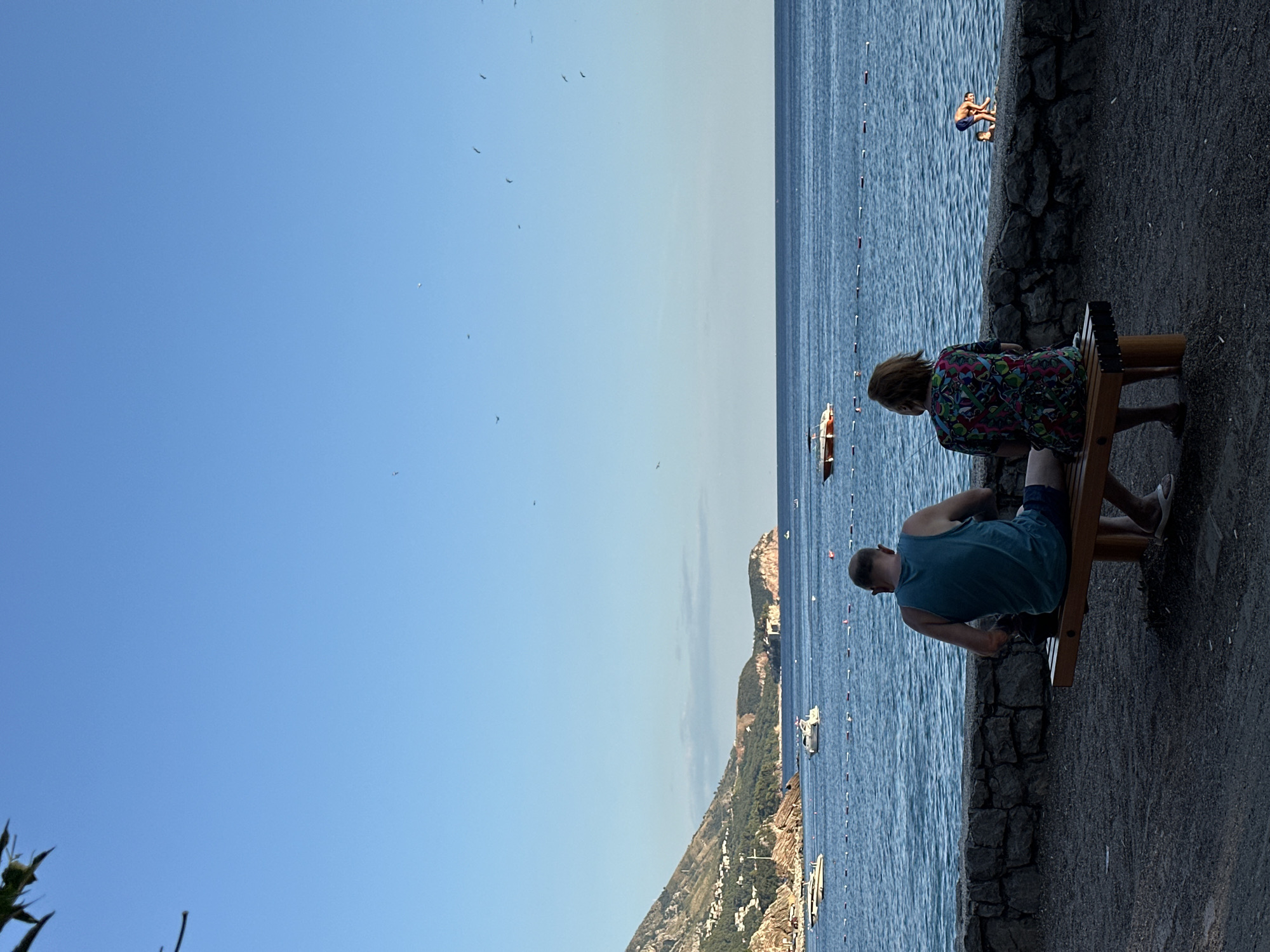 In the city of Boreti, Montenegro, two individuals sit on a wooden bench facing a calm sea under a clear blue sky. The man, on the left, wears a teal tank top and blue shorts, while the woman beside him sports a colorful patterned dress. Behind them, a stone wall separates the paved area from the beach. The sea is dotted with several boats and a floating line marker. In the distance, a mountainous coastline with scattered buildings stretches across the horizon. A small child is visible near the water's edge on the right side of the frame. Several birds are flying in the sky.