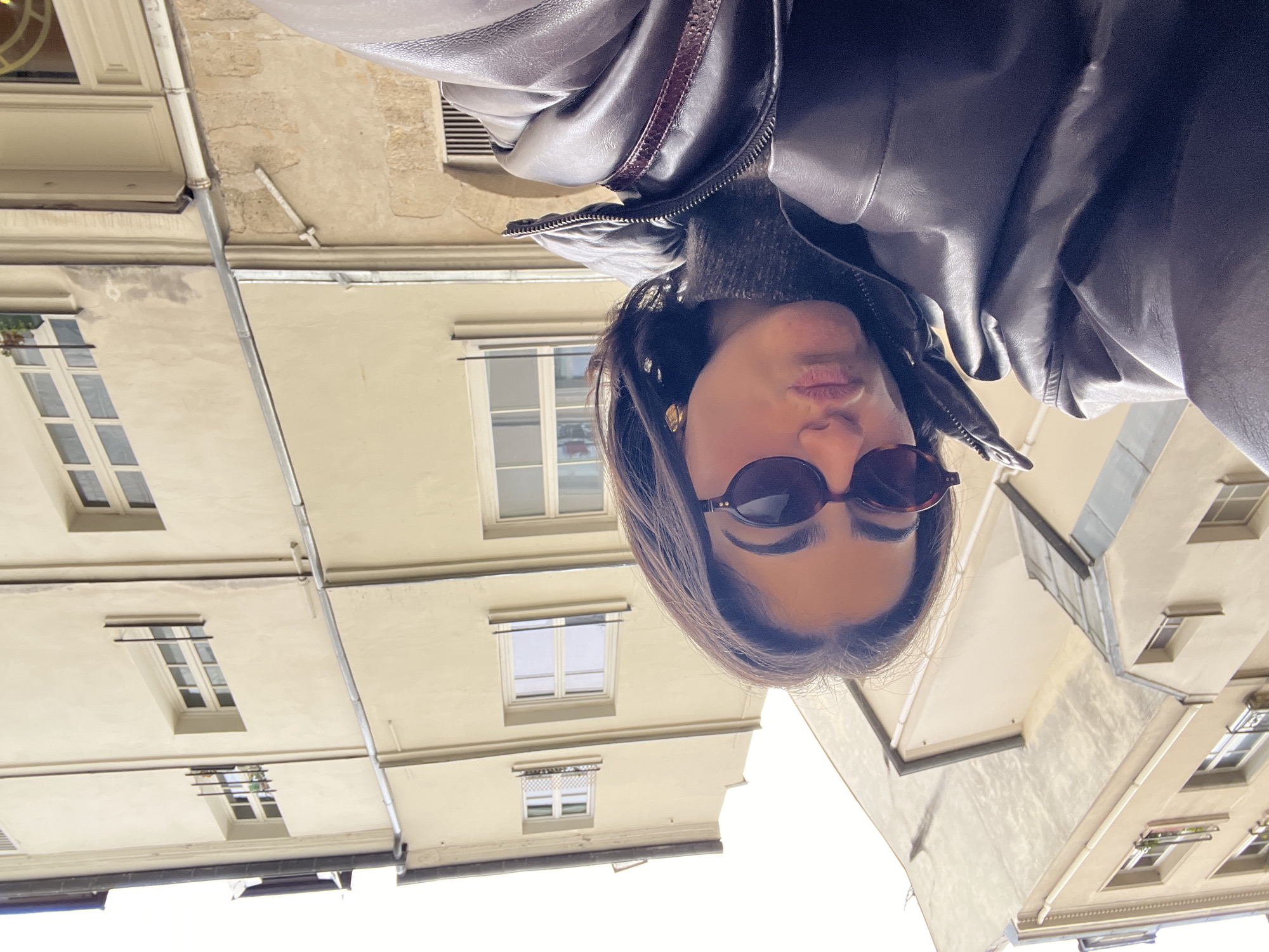 A low-angle shot features a person wearing a brown leather jacket and sunglasses in front of Parisian architecture. The buildings have light-colored facades with windows. A gutter is visible on one of the buildings. The sky is light and overcast.