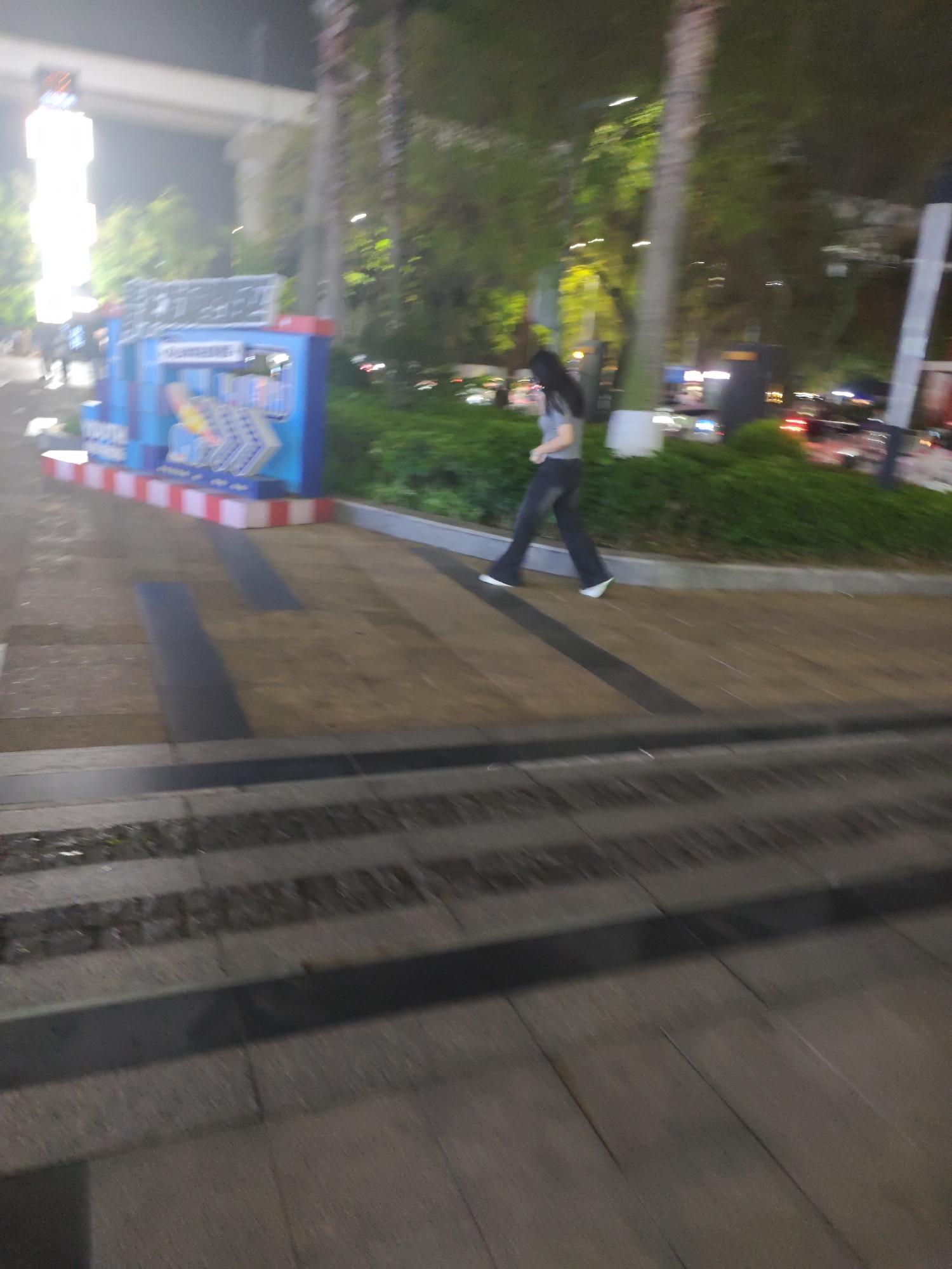 The image is a blurred, long-exposure shot taken at night in what appears to be a public area, possibly in the Chongqing High-tech Zone, China. It's difficult to ascertain definitive location details. The scene captures a woman in motion, walking from left to right, wearing dark pants and a light-colored top. She has long, dark hair. In the background, there are trees and buildings, blurred due to the motion and long exposure. A blue and red striped sign or structure is visible on the left. The foreground shows stone steps or paving, also blurred, indicating movement during the photograph's capture. There is ambient lighting from streetlights and buildings.