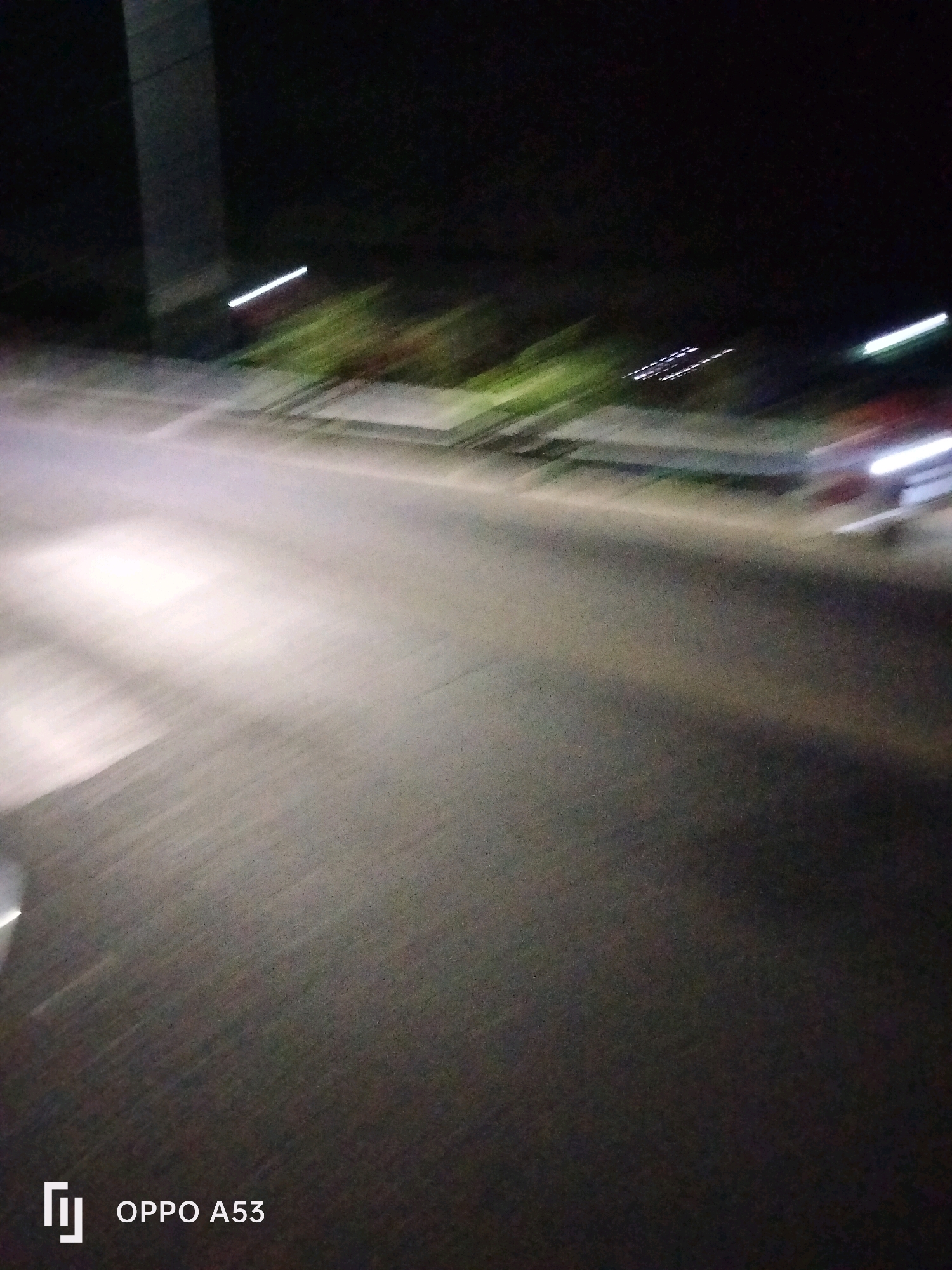 The image depicts a dark, blurry scene, likely taken at night while in motion. A road is visible, with headlights from a vehicle illuminating the surface and creating light streaks. In the background, there are blurred shapes that could be buildings or trees, with some lights indicating activity. The bottom left corner shows a logo that reads "OPPO A53," suggesting the image was captured with this model of phone. The overall impression is of a fast-moving vehicle on a road at night, possibly in a semi-urban or rural setting given the presence of what appear to be roadside elements. There is no specific information in the image to identify the location as Bendo, Indonesia.