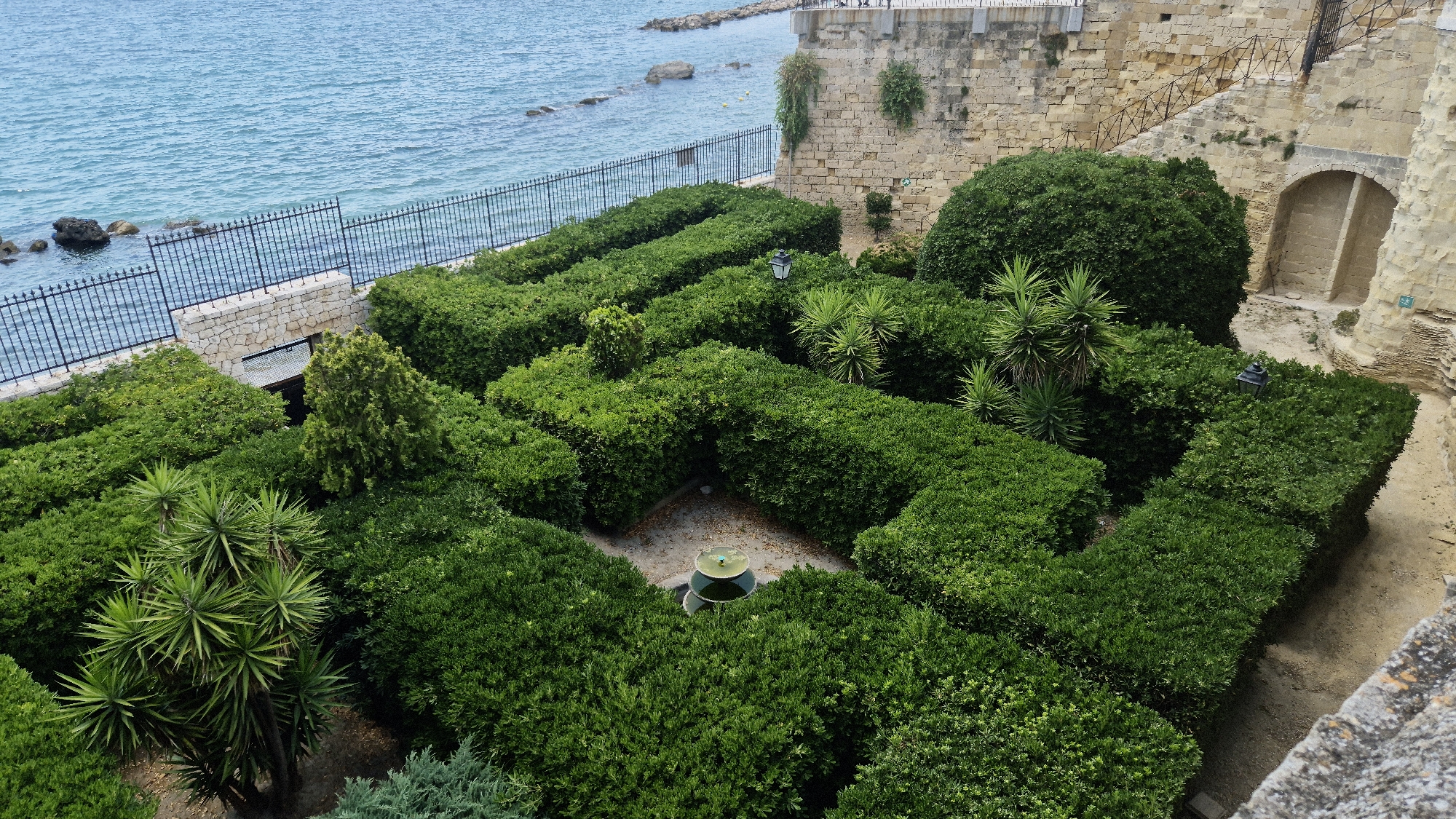 The image depicts a well-maintained hedge maze situated within a coastal fortress. The maze is densely planted with vibrant green bushes, forming intricate pathways and walls. In the center of the maze, a small circular fountain with a green-tinted water feature is visible. Scattered within the maze are a few spiky yucca plants and a single, conical evergreen tree. A traditional black lamppost stands at the edge of one of the maze's pathways.
Behind the maze, a weathered stone wall, characteristic of ancient fortifications, rises up. This wall is adorned with some creeping greenery. To the left of the maze, a black metal fence separates the garden from the blue, slightly choppy sea. The sea appears calm, with some rocks visible near the shore and a hint of a breakwater in the distance. The sky is overcast, suggesting a mild or cloudy day.
To the right of the maze, a sand-colored pathway leads towards an arched opening in the fortress wall, hinting at further exploration within the structure. The overall impression is one of a historical site integrated with a beautiful, formal garden. The time of day is indeterminate due to the overcast sky. No people are visible in the image. No text is visible. The location is identified as Taranto, Italy.