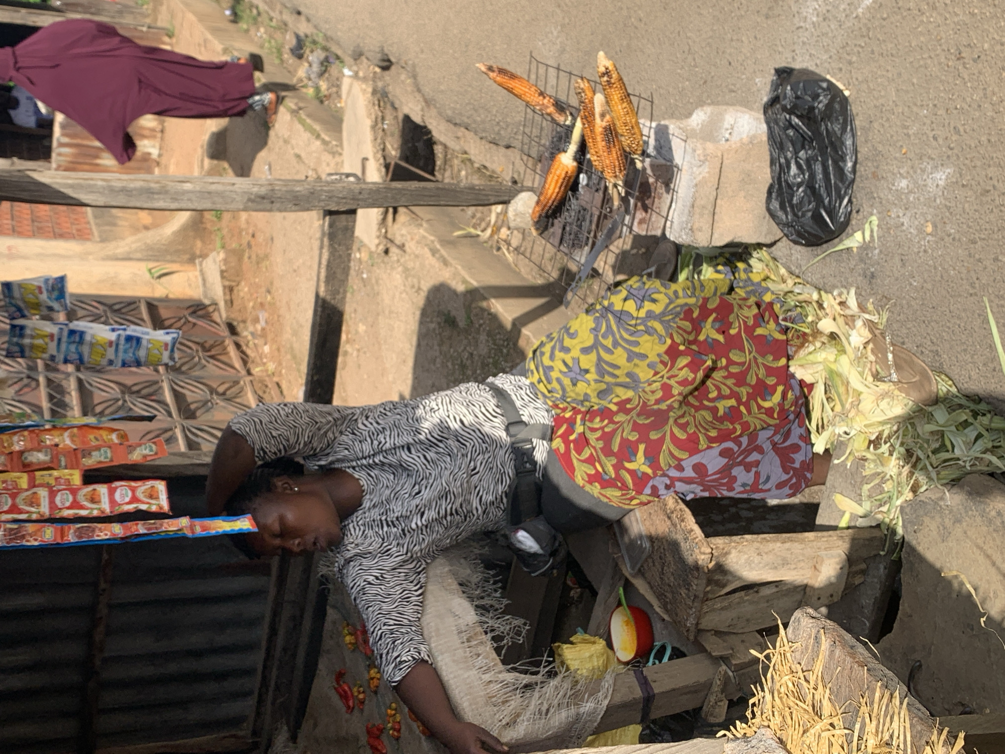 The image shows a woman selling roasted corn at a street-side stall in Jos, Nigeria. The woman is wearing a black and white patterned shirt and a colorful wrap skirt. She is seated behind a small wooden stall, with a charcoal grill in front of her, on which several ears of corn are roasting. To her left, on a wooden surface, are some red peppers and what appear to be small yellow fruits. Behind her, hanging from a structure, are numerous small packaged food items. In the background, a person in a dark purple hijab is walking away. The stall is set up next to a sidewalk, with a corrugated metal fence visible behind the stall. The ground is dusty and unpaved in some areas, with scattered debris. The overall scene depicts a typical bustling street market environment in Jos.