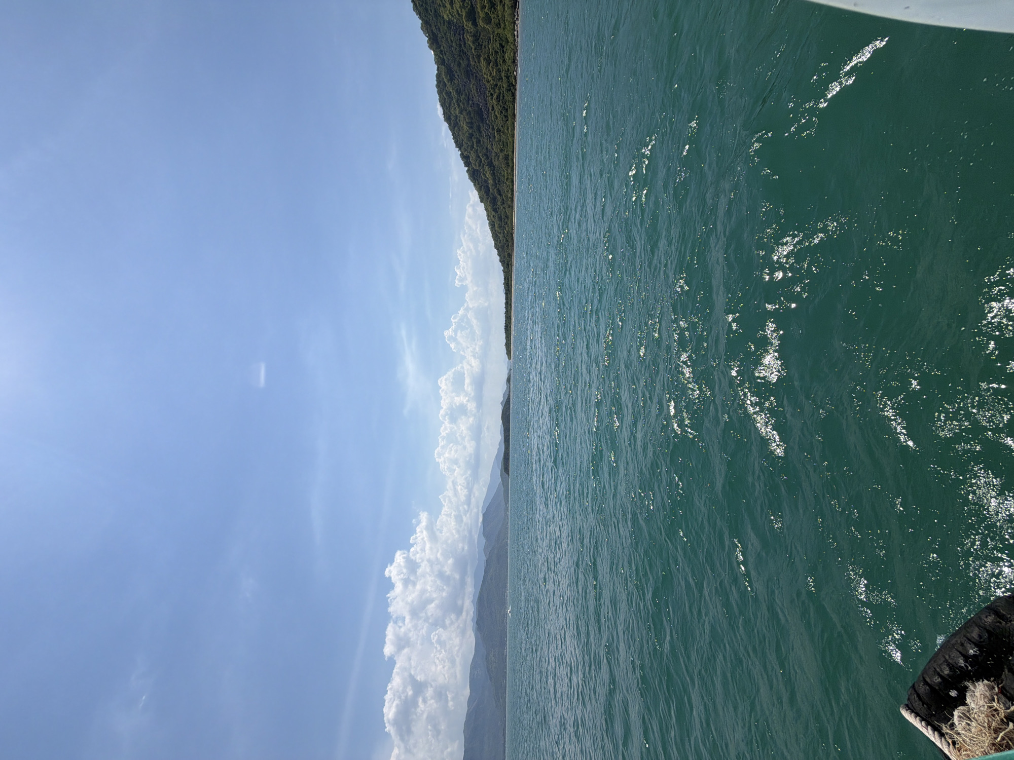 The image depicts a panoramic view of the sea in Bac Nha Trang Ward, Vietnam, on a clear day with scattered clouds. The turquoise water is calm, with gentle waves reflecting the sunlight. In the distance, verdant, tree-covered hills form a dramatic coastline. The sky is a brilliant blue, with fluffy white clouds adding texture and depth. The perspective suggests the photograph was taken from a boat, with the edge of the vessel visible in the lower left and right corners. There are no people or specific structures visible in the scene, emphasizing the natural beauty of the coastal landscape. The overall impression is one of tranquility and natural splendor.