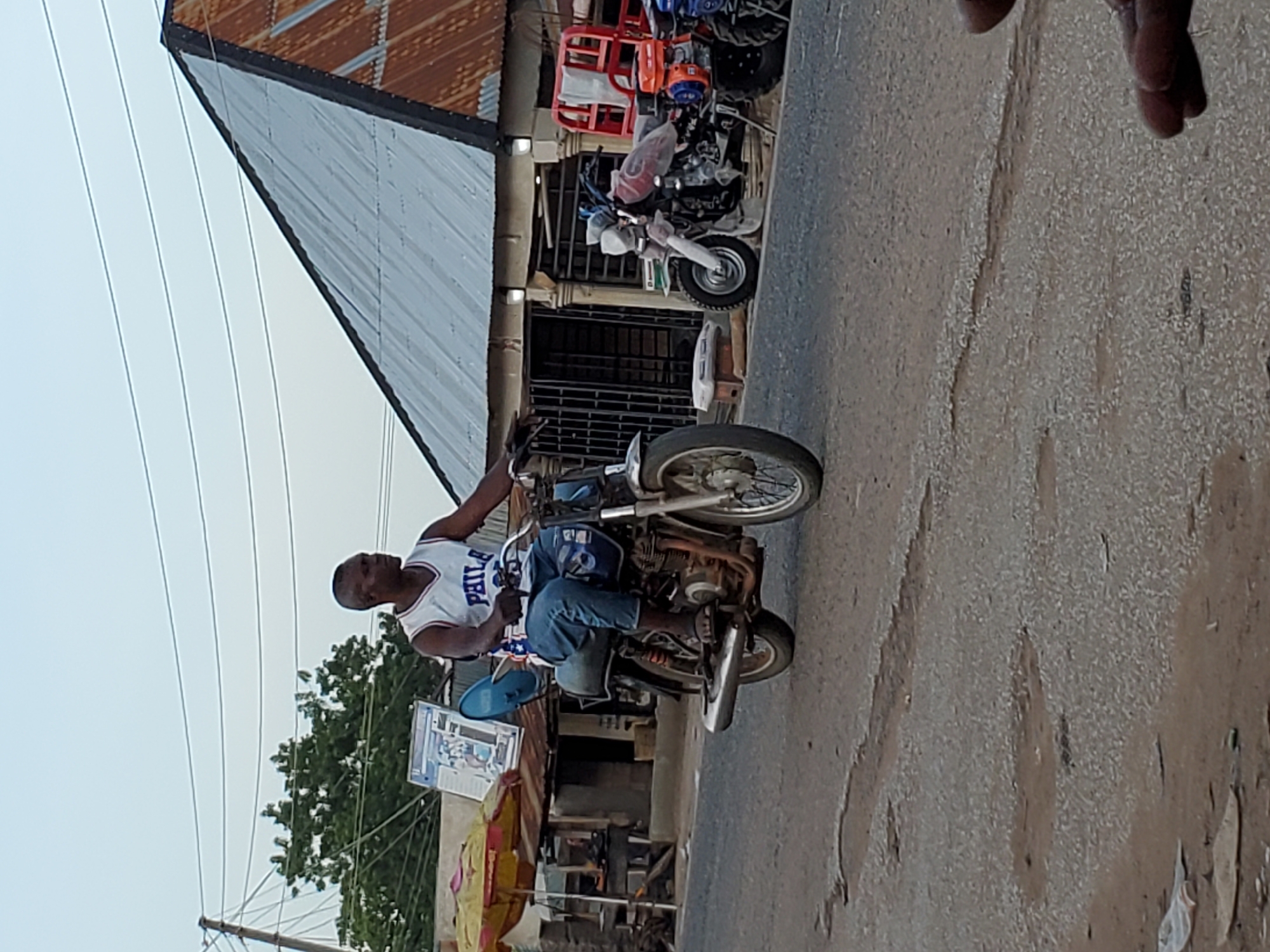An eye-level shot captures a man riding a motorcycle on a dusty or unpaved street in Mararaba, Nigeria. The image is rotated 90 degrees clockwise, so the man appears to be riding upwards relative to the frame.
The central subject is a man of African descent, wearing a white sleeveless tank top with "PHILA" printed in blue text across the chest, and blue jeans. He is seated on a dark-colored motorcycle, steering it with both hands. A rectangular poster or sign, featuring images and text, is affixed to the front of his motorcycle, partially obscuring the front wheel.
In the background, on the right side of the image (when oriented correctly), a building with a corrugated metal roof is visible. Several other motorcycles are parked near this building, suggesting a street with commercial activity or residential garages. One of the parked motorcycles has a bright red cargo rack on its rear. Above the street and buildings, power lines crisscross against a light, clear sky. Green foliage from trees is visible in the distance on the left side. The road surface is visibly uneven and sandy/dusty. The lighting suggests it is daytime, possibly morning or late afternoon, given the soft, diffused light.