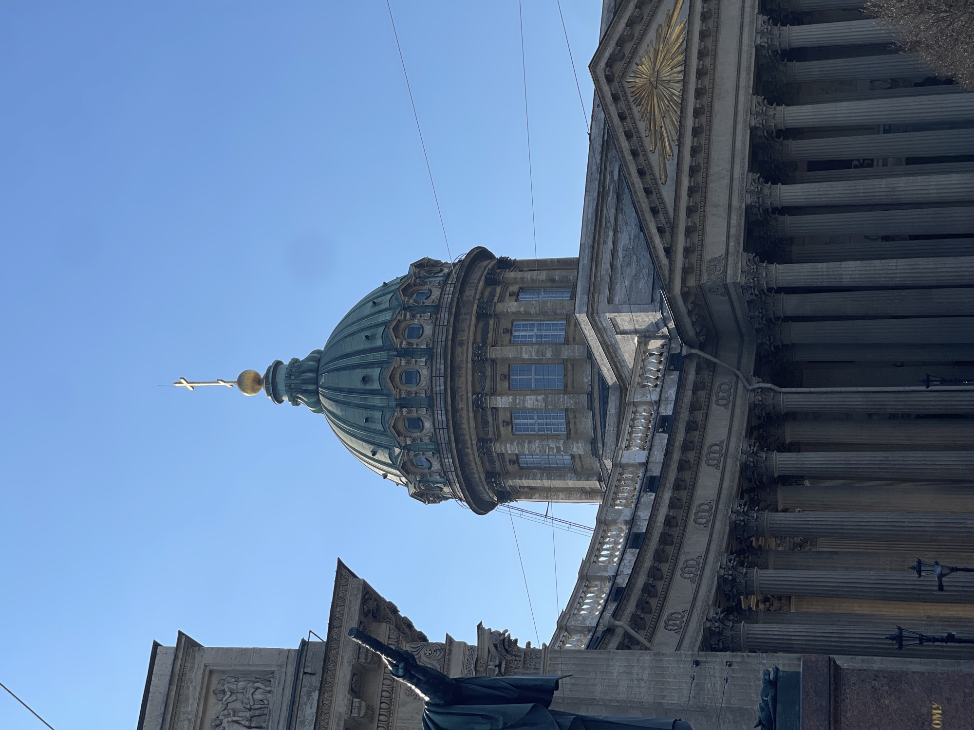 The image shows the Kazan Cathedral in Saint Petersburg, Russia, viewed from below. The focus is on the cathedral's dome, topped with a golden cross, and the adjacent architectural details, including columns and ornamentation. Below the cathedral, a statue is partially visible, adding to the perspective and depth of the shot. The sky is clear and blue. The image appears to be taken during the day.