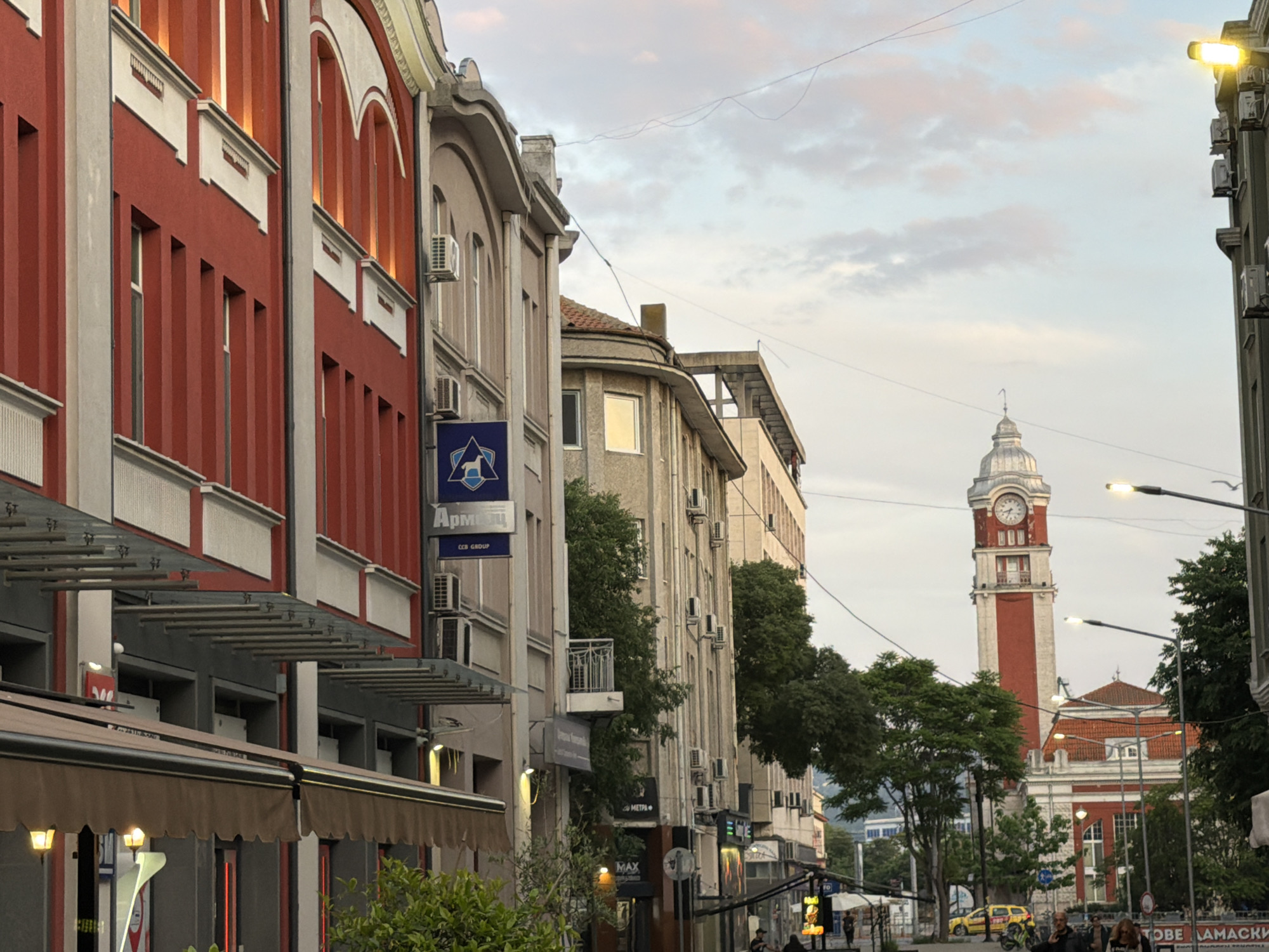 This image captures a vibrant street scene in what appears to be a European city during late afternoon or early evening. The sky, partially cloudy with soft grey and pink hues, suggests the approach of dusk, with some artificial lights already glowing.
On the left, a multi-story building with a striking red facade and white architectural details, including arched windows and decorative elements, dominates the foreground. Below it are brown awnings extending over what might be shops or cafes. Adjacent to it, another building is light beige with arched windows and numerous external air conditioning units. A prominent blue sign on this building displays a white horse/dog logo and the Cyrillic text "Армейц" (Armeets), with "CCB GROUP" written below.
Further down the street, various other buildings line the thoroughfare, some featuring balconies and more air conditioning units. Lush green trees are interspersed among the buildings, providing some natural greenery. Overhead utility wires crisscross the sky.
In the distance, the focal point is a tall, distinctive clock tower, primarily red and white, with a large clock face visible near its top. The tower is crowned with a domed structure. Below the clock tower, part of a building with a red roof and white columns can be seen.
The street itself appears busy, with several pedestrians visible on the sidewalk in the lower right, though their features are indistinct. A yellow car, possibly a taxi, and some motorcycles are parked further down the street near the clock tower. Streetlights are visible, some emitting a warm yellow glow. Other visible text includes "MAX" on a distant sign and partial Cyrillic text "ТОВЕ АМАСКІ" or similar on a sign in the far lower right.