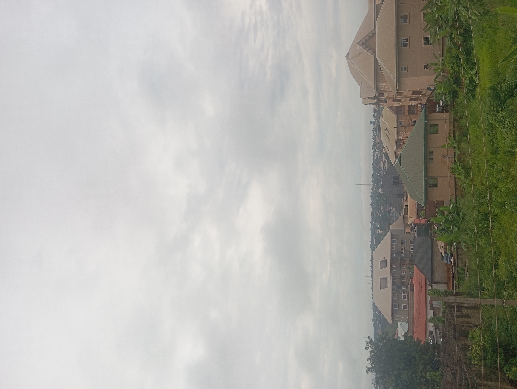 A vertical shot shows an aerial view of Nkpor, Nigeria, on a cloudy day. Several multi-story residential buildings with light brown, beige, and green roofing are visible. Banana trees and lush green vegetation surround the buildings. Distant buildings in the city fade into the hazy background, culminating with a cloudy sky. Power lines are visible in the foreground.