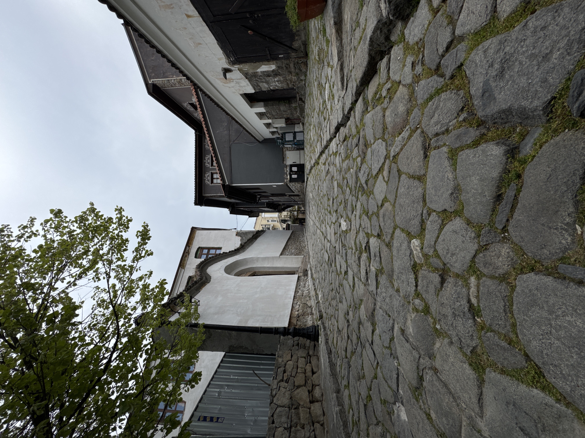 The image shows a street in the Old Town of Plovdiv, Bulgaria, characterized by cobblestone pavement. Buildings line either side of the street, with architectural details including exposed stone foundations, white stucco walls with decorative flourishes, and dark wood trim. A tree with green leaves is visible on the left. The sky is overcast.