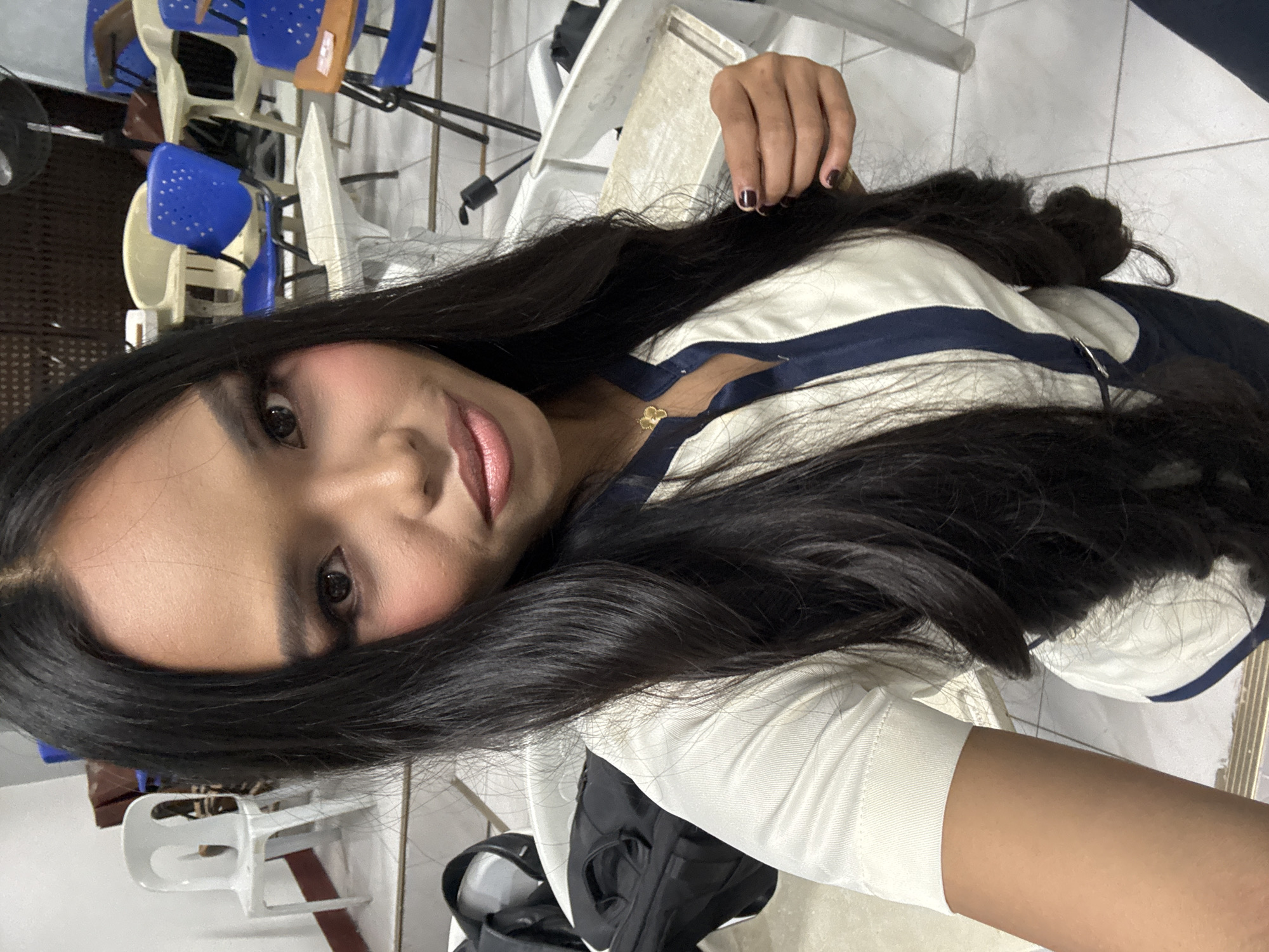 In a room with tiled flooring, a young woman with long, dark hair and makeup poses for a selfie. She wears a white collared shirt with navy blue trim and a navy blue skirt. A gold necklace with a flower pendant adorns her neck. In the background, various classroom chairs and tables are visible. One white plastic chair is positioned to the left of the frame. The scene appears to be indoors, possibly in a school or office environment within Davao City, Philippines.