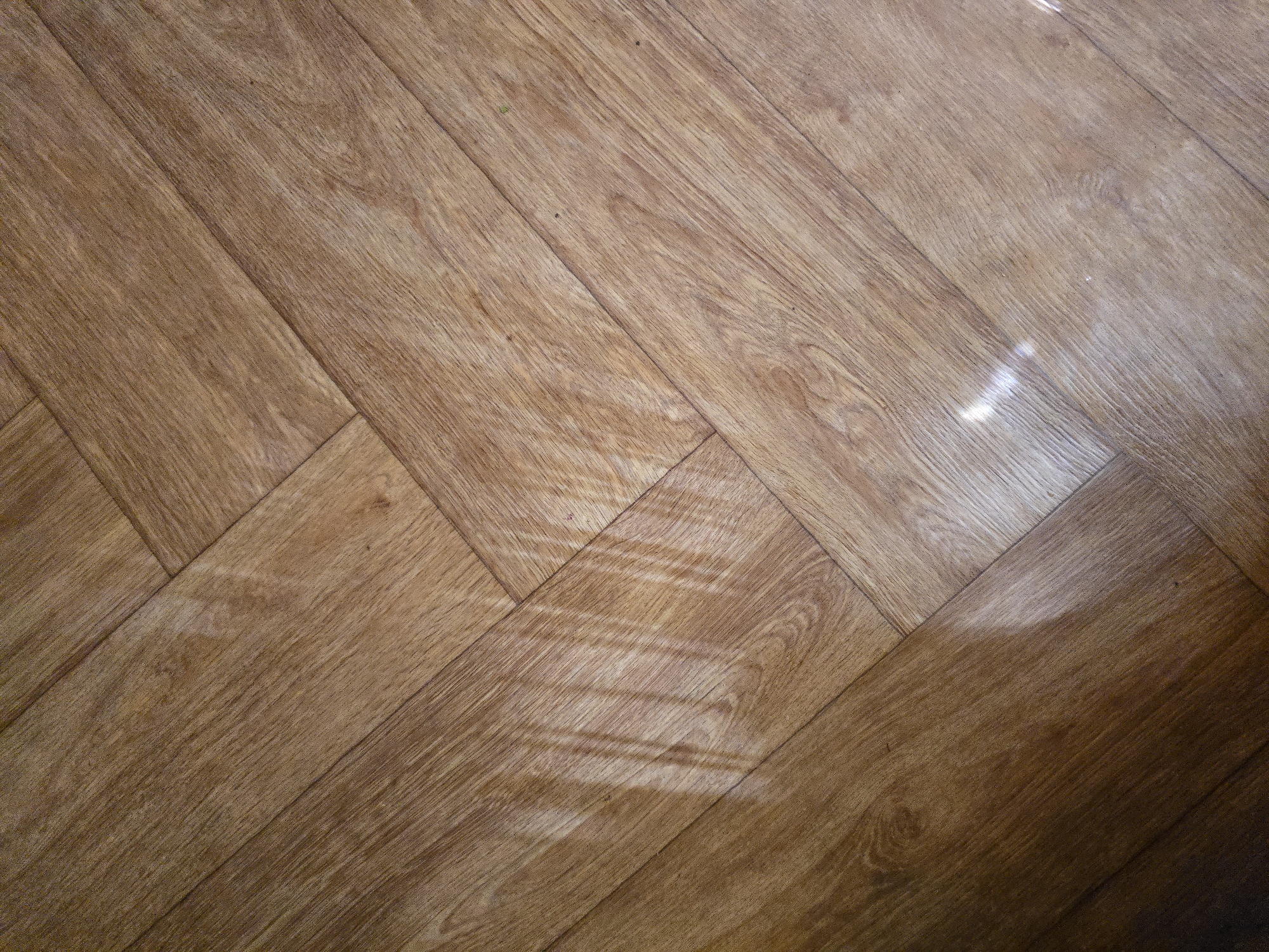 The image shows a close-up, high-angle view of a light brown wooden floor laid in a herringbone pattern. The floorboards are narrow and elongated, with visible wood grain and some lighter, streaky markings. There are reflections of light on the floor, particularly a prominent streak of light running diagonally across the left side of the image, creating a striped effect. The lighting suggests an indoor environment. No people, animals, or other objects are visible. The location is Unknown, Unknown.