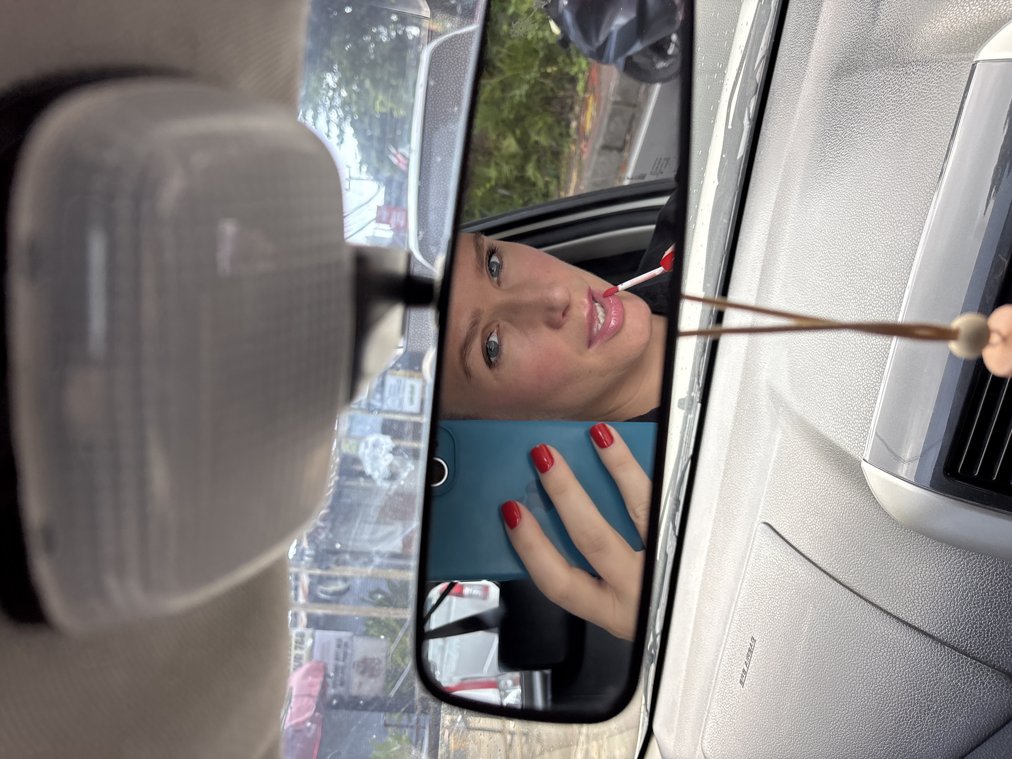 The image provides an interior view of a car, primarily focused on the rearview mirror. Reflected in the mirror is a woman in the process of applying lip gloss or lipstick to her plump lips. Her left hand, adorned with bright red nail polish, holds a teal-colored smartphone which is pointed towards her, suggesting she is taking a photo or video of herself in the mirror. The woman has light-colored eyes and is looking directly into the reflection.
Around the rearview mirror, parts of the car's interior are visible, including the textured gray dashboard and an overhead light fixture. A small, decorative charm with wooden beads hangs near the mirror on the right side. Faintly visible on the dashboard is the text "SRS AIRBAG".
The rearview mirror also reflects the scene outside the car through the front windshield. This exterior view shows a street with green foliage, a building, and possibly other vehicles, indicating a daytime setting. The overall image is rotated approximately 90 degrees clockwise, causing the woman's head to be tilted and the exterior view to appear on its side. The location context is Kuta Selatan, Indonesia.
