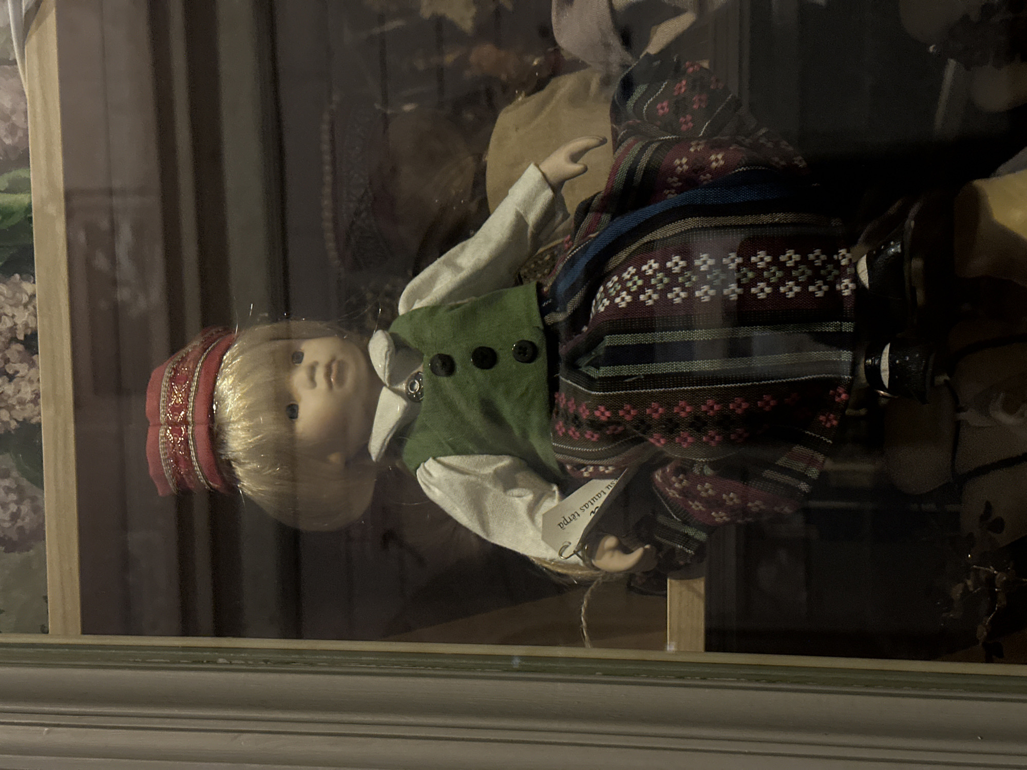 The image shows a doll dressed in traditional Latvian clothing, likely a souvenir or part of a cultural display. The doll is positioned behind glass, possibly in a shop window or museum display case. Based on the traditional clothing, this is most likely in Riga, Latvia. The doll wears a red head covering, a green vest, and a patterned skirt.