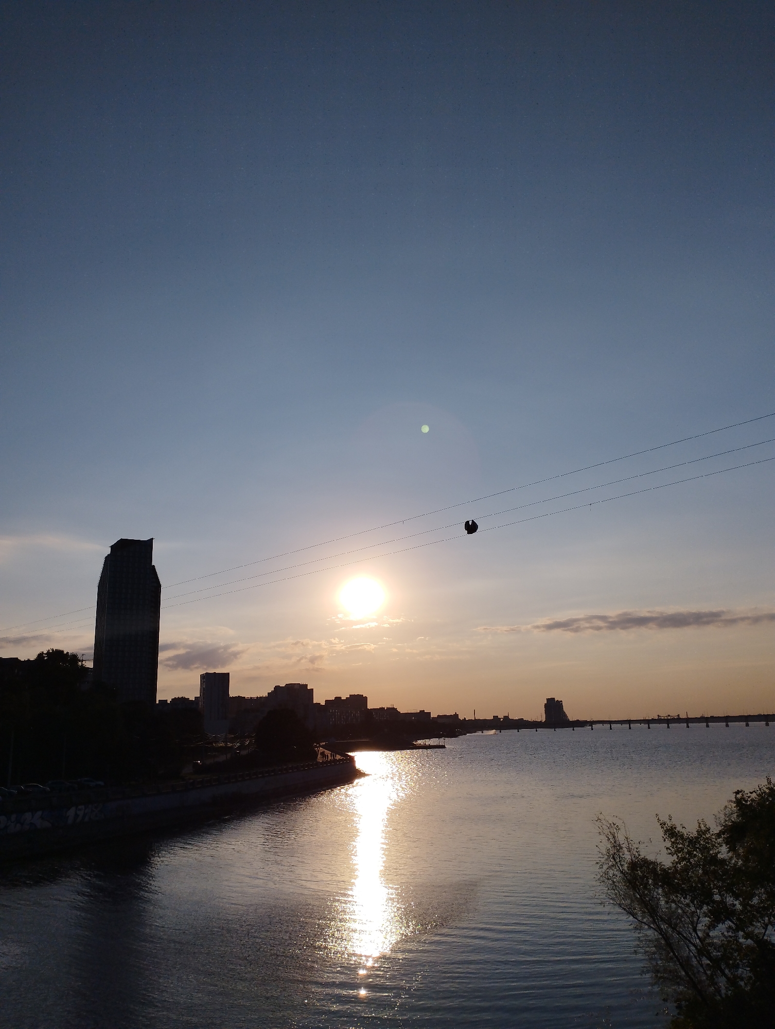 This is a vertical, silhouetted image of a city skyline along a body of water at sunset. The sun is low in the sky, casting a bright reflection on the water and creating a hazy glow. Several buildings of varying heights are visible along the shoreline, including a tall, slender skyscraper on the left. In the distance, a bridge spans the water. Two thin wires stretch across the sky, with a dark, round object hanging from one of them. The sky is a gradient of blue and orange, with a few wispy clouds. The overall scene is serene and somewhat melancholic, with the fading light and the quiet cityscape.