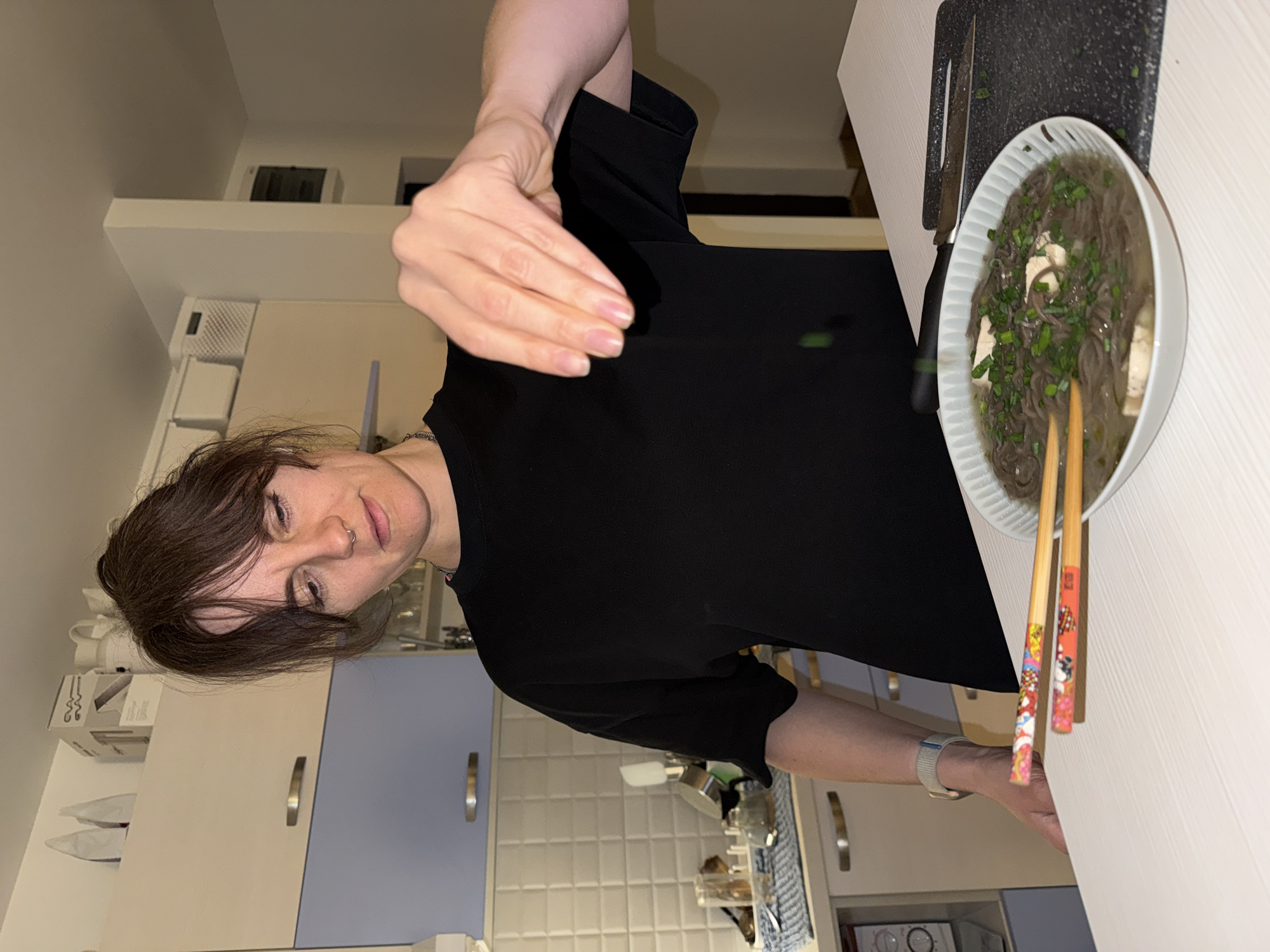 A woman in a black shirt is adding greens to a bowl of soup. The soup contains noodles, tofu, and other greens. Chopsticks are resting on the bowl. A cutting board and knife are next to the bowl. The scene appears to be a kitchen.