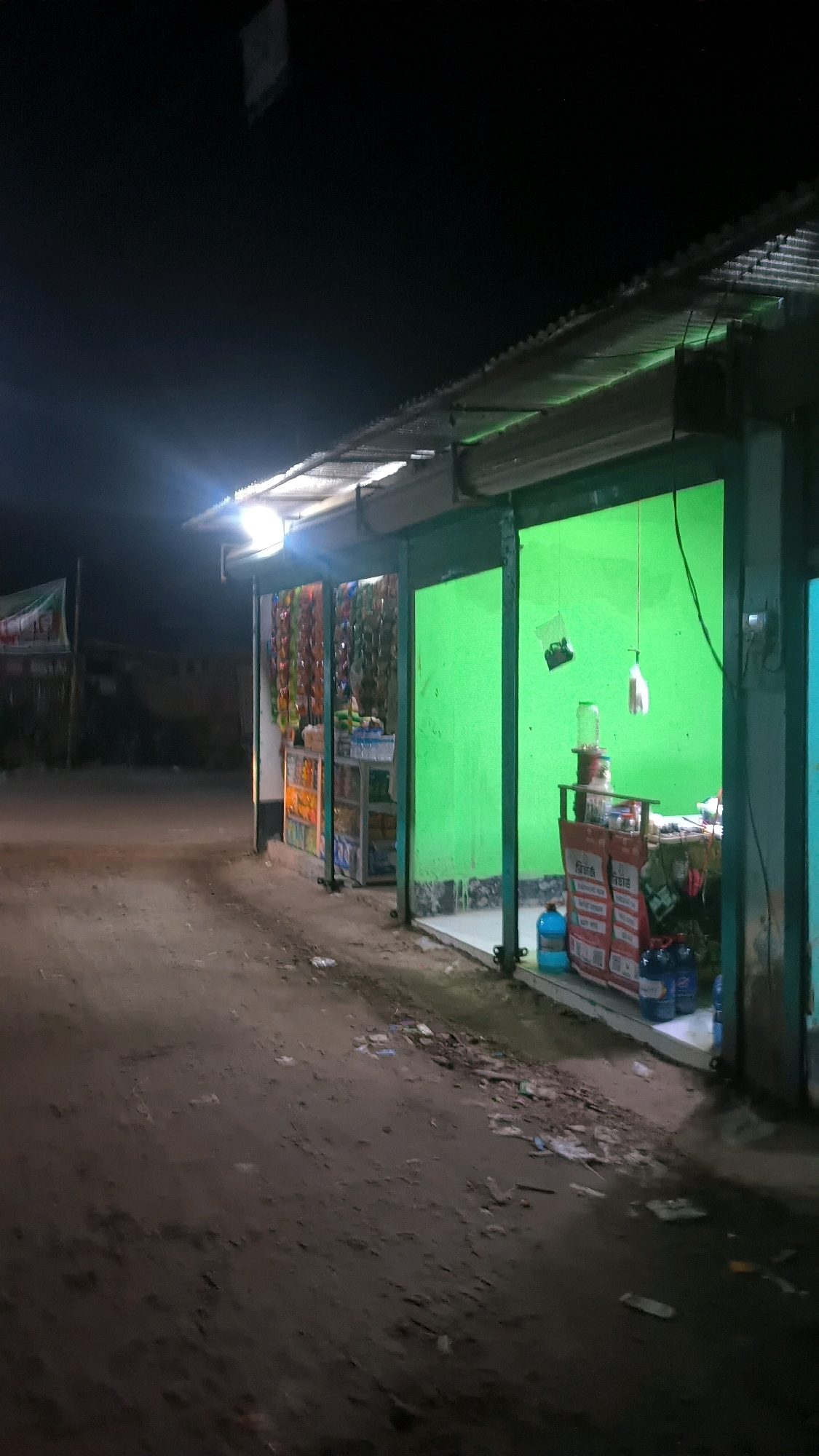 The image shows a small, open-air storefront at night in Begumganj Upazila, Bangladesh. The storefront is lit by a bright overhead light. The front of the store features a green wall and an open entrance where shelves of goods are visible. Packaged items, such as snacks and drinks, are displayed on the shelves. A water cooler jug is placed on a counter inside the store, along with other merchandise. There are blue plastic bottles of liquid outside the store. The ground in front of the store is dirt and appears littered with debris. A road or path runs alongside the storefront. The overall impression is that of a local convenience store or small shop operating at night.