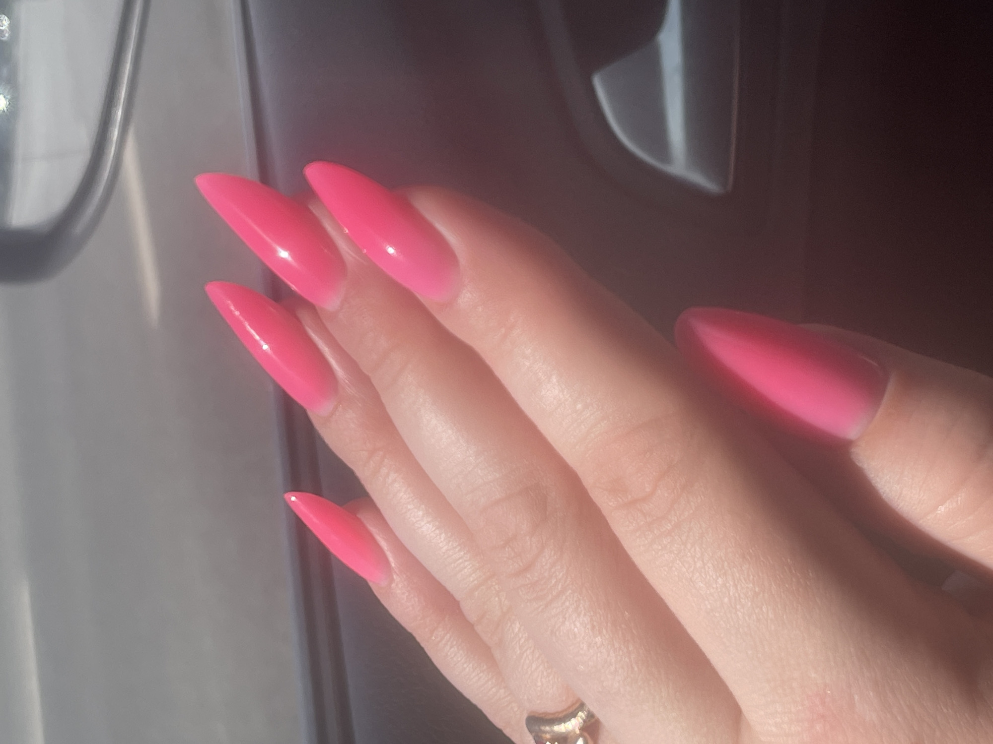The image shows a close-up view of a person's hand with bright pink, stiletto-shaped acrylic nails. The nails have a gradient effect, transitioning from a lighter pink at the base to a brighter, more saturated pink at the tips. A thin, gold-toned ring with a knot design is visible on the ring finger. The hand is resting against a dark, textured surface, possibly the interior of a car. The lighting suggests it is daytime, with sunlight casting shadows. There is no information in the image that could identify the location as Samara, Russia.