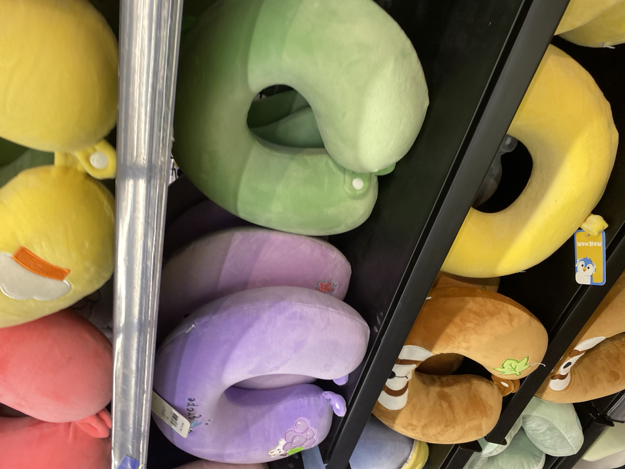 This is a close-up, eye-level shot of a retail display of plush neck pillows. The pillows are arranged on black shelves. In the top row, partially visible, are pink and yellow pillows. The middle row features several U-shaped neck pillows, with a prominent lavender one on the left, a pink one next to it, and a green one on the right. The bottom row shows more neck pillows, including a brown one with a sloth design and a bright yellow donut-shaped pillow. The lighting is bright and appears to be from overhead store lights. The image is focused on the products, with no people or other environmental details visible. The scene is likely in a store or market in Banda Aceh, Indonesia, given the context provided.