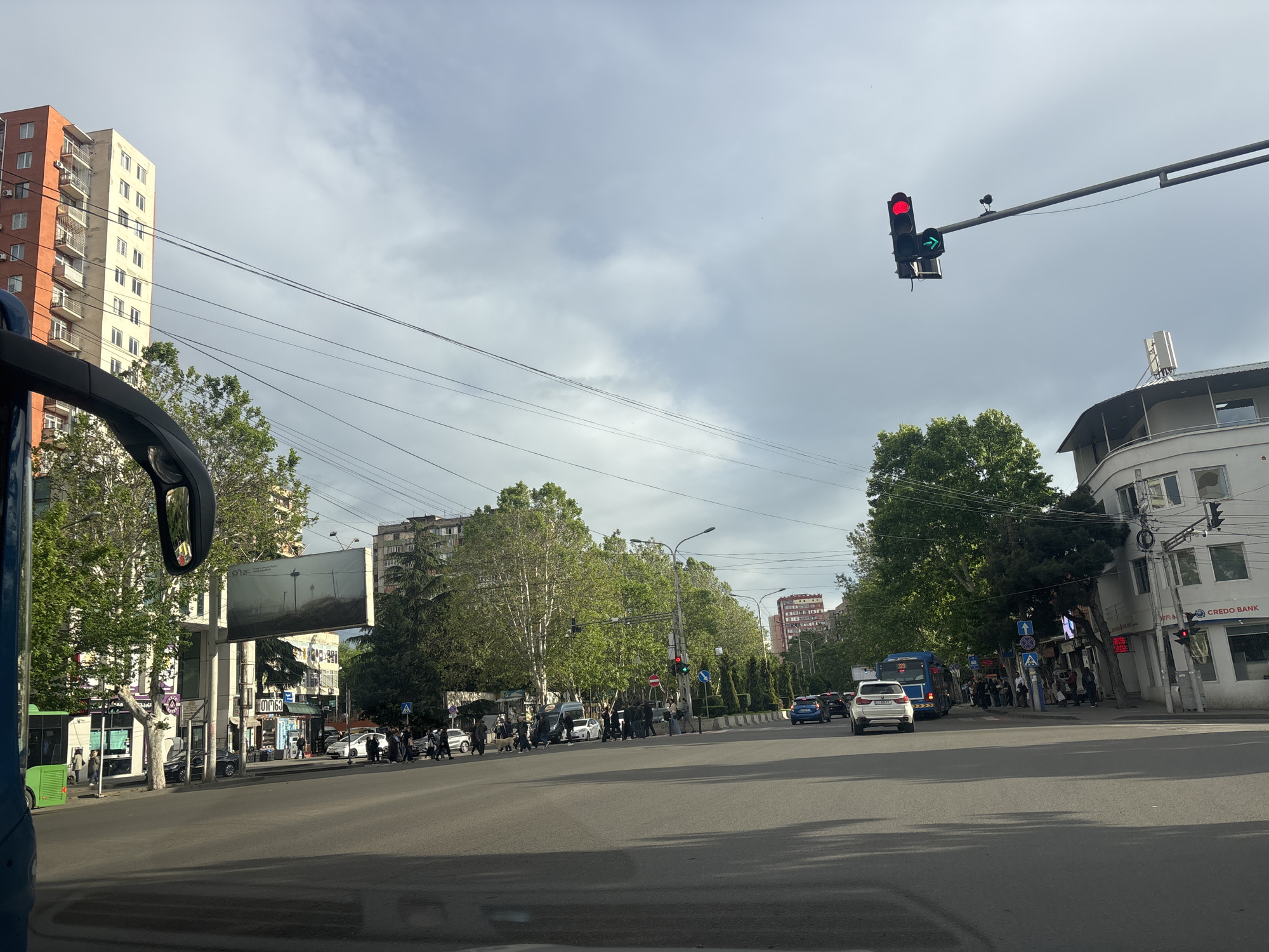 The image shows a street scene in Tbilisi, Georgia. It is daytime, and the sky is overcast. The view is from inside a vehicle, likely a bus or car, looking out onto an intersection. The road is paved and appears wet in the foreground, reflecting light. In the distance, people are crossing the street at a crosswalk. Several vehicles are on the road, including cars and buses. Buildings of varying heights line the street, including an orange and white apartment building on the left, and a white building with the sign "CREDO BANK" on the right. Streetlights and traffic signals are visible, with a traffic light showing a red light with a green arrow to the right. Trees are scattered throughout the scene, adding greenery to the urban environment. A large billboard is on the left side of the intersection. The general atmosphere is urban, with a mix of residential and commercial buildings, pedestrian activity, and vehicular traffic.