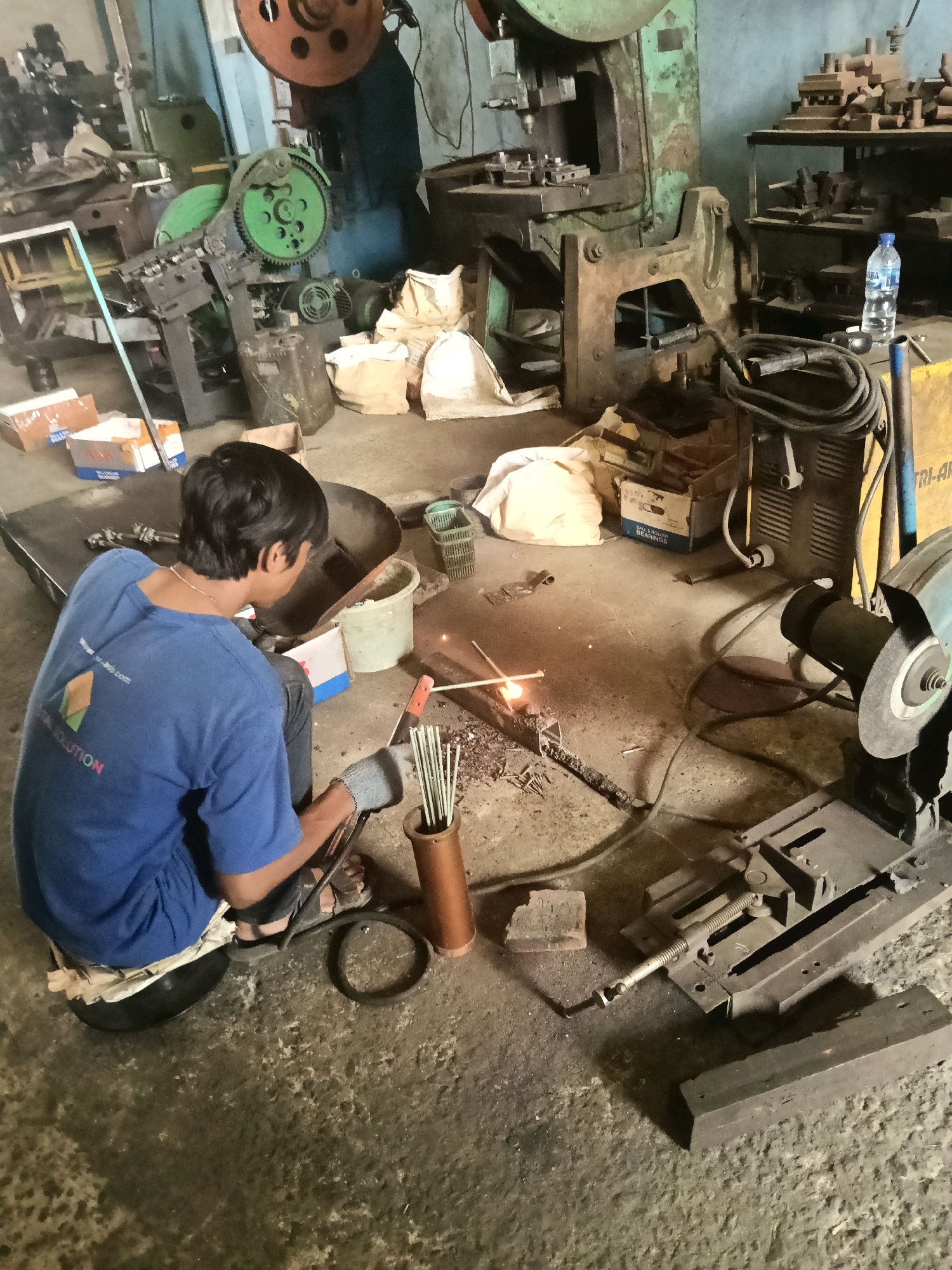 In Belimbing, Indonesia, an individual is welding in a dimly lit workshop. The person, wearing a blue t-shirt, dark pants, and gloves, is seated on a black cushion on the floor, focused on the welding task at hand. Sparks fly from the welding rod, illuminating the immediate area. Surrounding the worker are various pieces of industrial equipment, including a large green press, a cutting saw, and metal scraps. Bags of materials and boxes are scattered around the floor, suggesting an active workspace. A bottle of water and a metal bucket are also visible. The environment appears to be a working industrial space, possibly a metal fabrication shop.