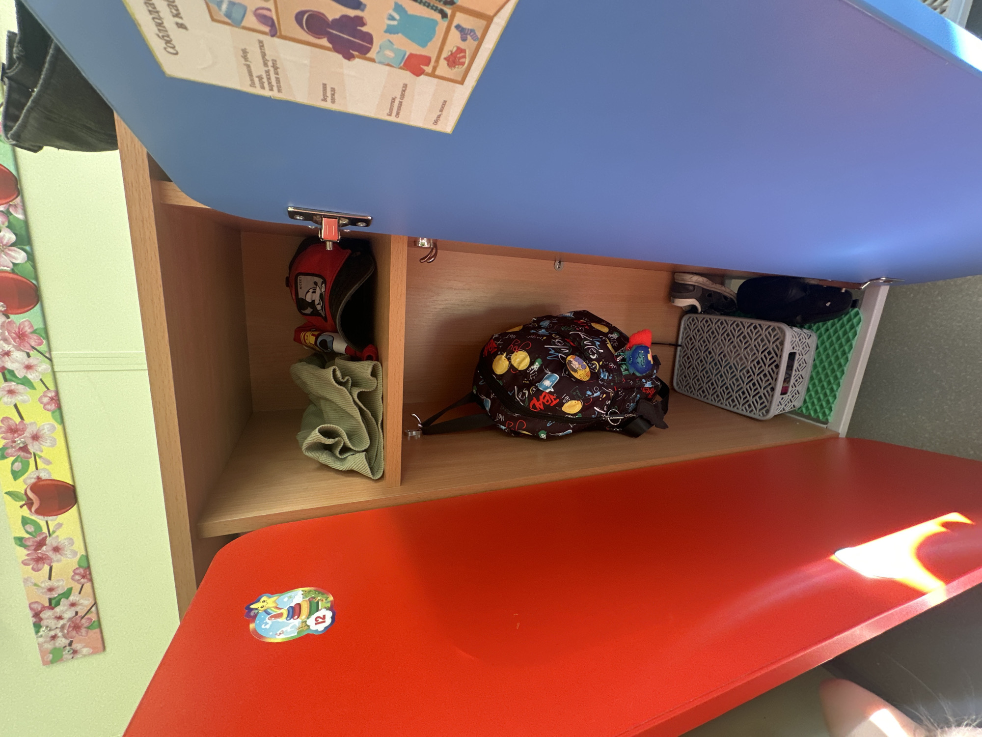 This image depicts the interior of a locker, likely in a school or daycare, as evidenced by the cartoon decorations on the doors and the organized storage. The locker is divided into compartments. On the top shelf of the left compartment, a green folded cloth is visible. Below that, a patterned backpack hangs from a hook. To the right of the backpack, on the floor of the compartment, is a gray basket with a geometric pattern, and below that, a pair of black shoes. To the right of the backpack, on the top shelf of the right compartment, is a red baseball cap with a Mickey Mouse logo and a red toy. A blue door of an adjacent locker is visible on the right side of the image, with a poster displaying illustrations of clothing items and their categories. The locker doors are red on the left and blue on the right. The overall environment appears to be indoors with a light green wall in the background. The specific location in Novoaltaysk, Russia, cannot be determined from the visual information provided in the image.