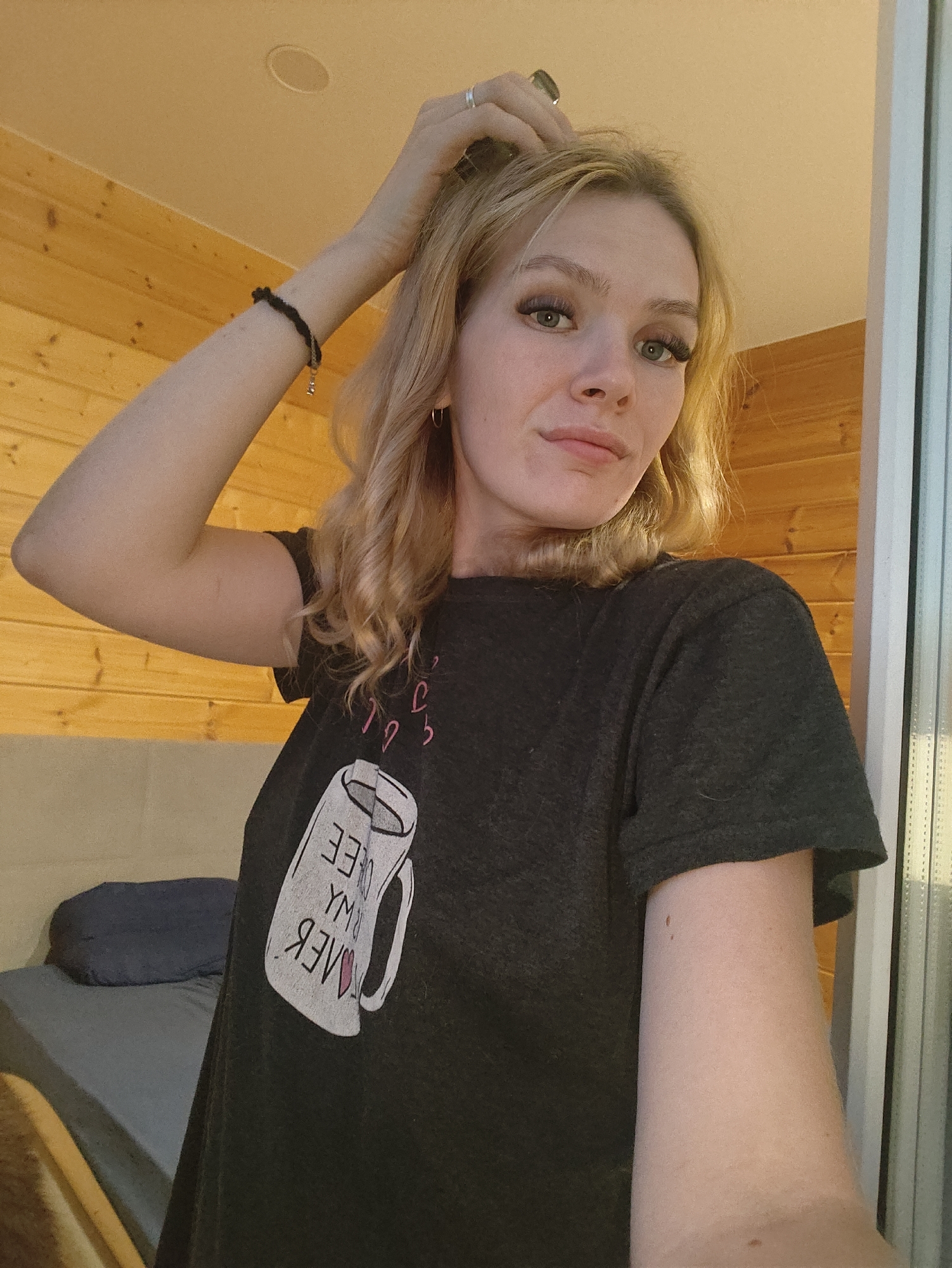A young woman with blonde, wavy hair and light blue eyes is taking a selfie. She is wearing a dark gray t-shirt with a graphic of a mug that says "COFFEE MY WAY" in reverse. Her left arm is raised, touching her hair, and she is wearing a black beaded bracelet with a small charm and a ring on her right index finger. She is positioned in front of a wooden-paneled wall. To the left of the woman, a blue pillow rests on a bed. The overall setting appears to be an indoor space, possibly a bedroom. The image does not contain any specific landmarks or visual cues to indicate the location within Русский Вожой, Russia.