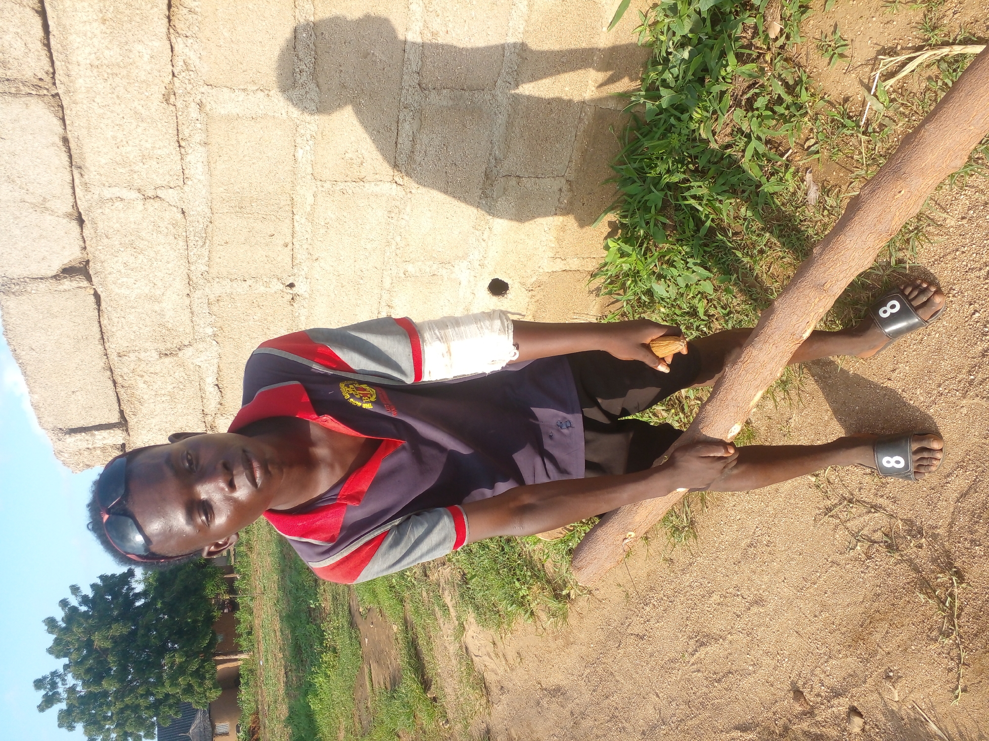 A young Black male is pictured outdoors, holding a large piece of wood. He is wearing a blue and red polo shirt, black shorts, and black sandals with the number 8 on them. His left arm is bandaged. He is standing on a dirt path next to a wall made of concrete blocks. There are green plants growing in front of the wall, and a tree is visible in the background. The sky is blue and clear. The scene appears to be in a rural or undeveloped area.