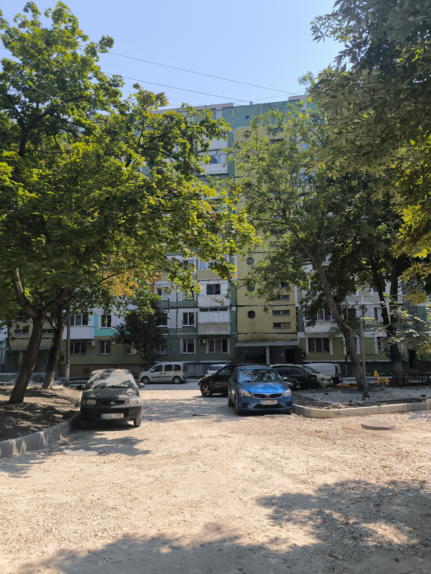 The image depicts a street scene in Chișinău, Moldova, under a clear blue sky on a sunny day. In the foreground, a dirt and gravel-covered area serves as parking. Several cars are parked here, including a dark-colored sedan on the left and a bright blue sedan on the right, both facing forward. Behind the parking area, a multi-story apartment building dominates the background. The building has a colorful facade, with sections painted in shades of yellow, green, and blue. Numerous windows and balconies are visible on the building's exterior. Lush green trees with dense foliage are scattered throughout the scene, providing shade and framing the apartment building. The trees are in full leaf, indicating a warm season. In the middle ground, more vehicles are visible, including a white van and other cars. The overall impression is one of an urban residential area.