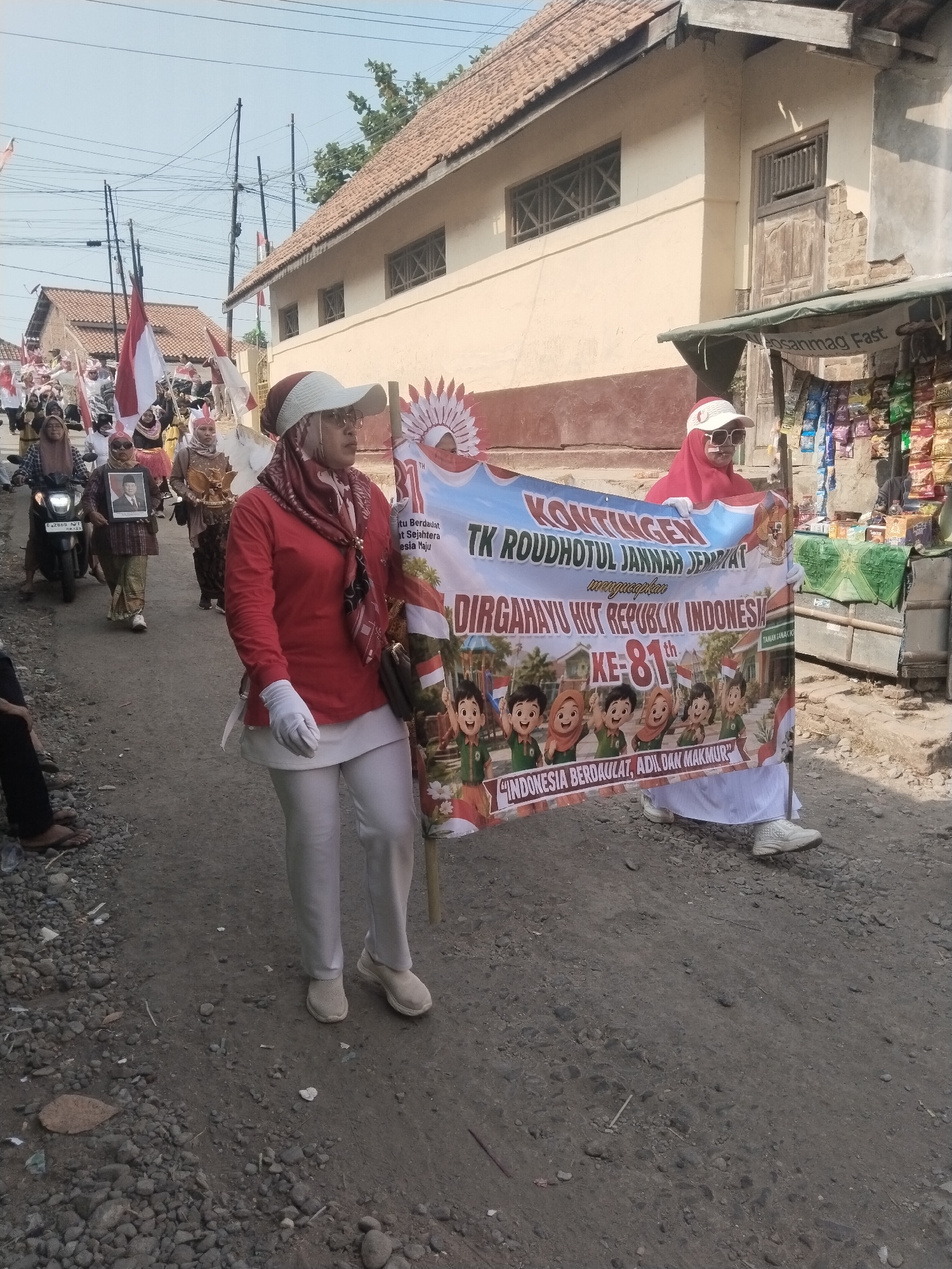 A parade is being held on a dirt road in Jembayat, Indonesia. A group of people, mostly women wearing headscarves, are marching. The women are carrying flags and a banner that reads "KONTINGEN TK ROUDHOTUL JANNAH JEMBAT, DIRGAHAYU HUT REPUBLIK INDONESIA KE-81 TH" and "INDONESIA BERDAULAT, ADIL, DAN MAKMUR". One woman in the foreground is wearing a red long-sleeved shirt, white pants, a red headscarf, and sunglasses, and is holding the banner. The buildings in the background appear to be simple, one-story structures with tiled roofs. There is a small shop on the right side of the road selling snacks and drinks. The sky is clear and the sun is shining.